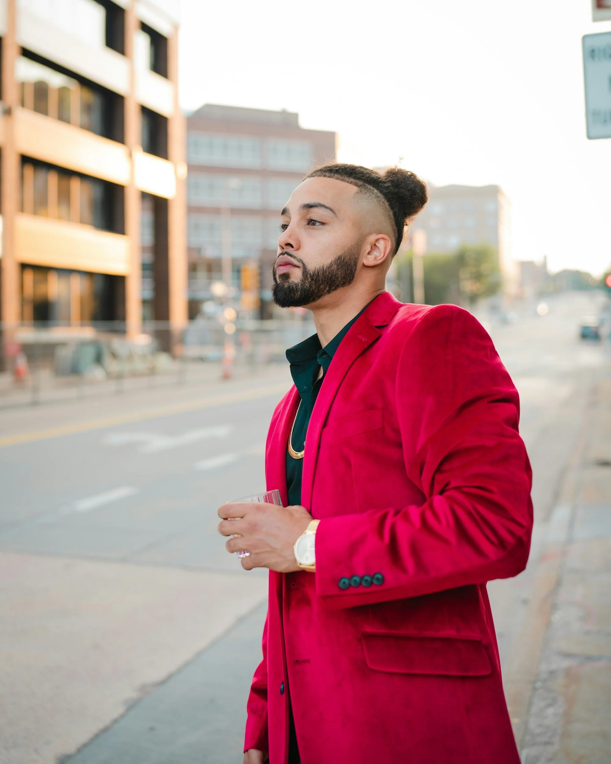 A man with a meticulously styled beard and a top knot hairstyle, wearing a red blazer and holding a drink, stands on a city street with modern buildings and traffic in the background.