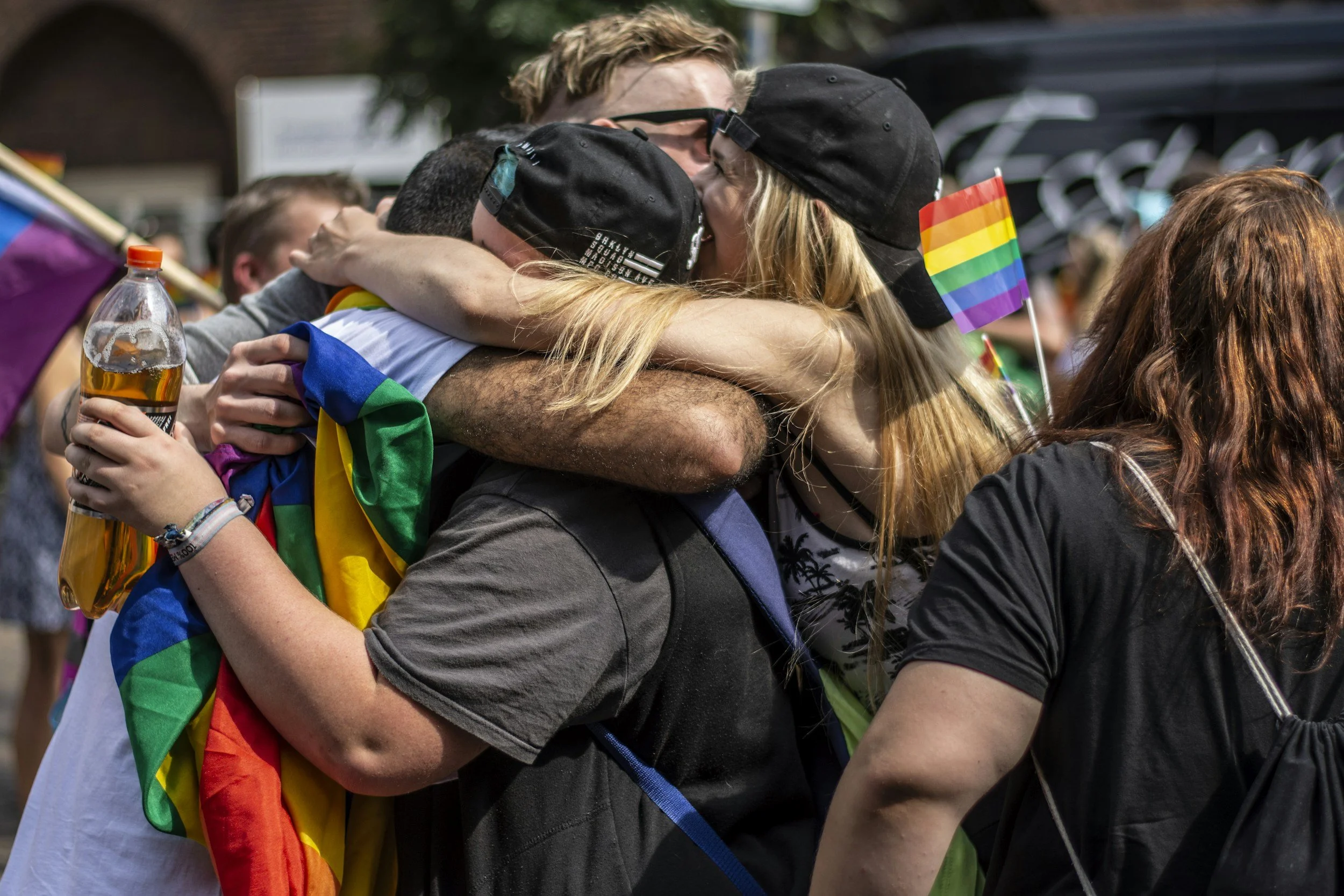 Two people hugging at a pride event, holding small rainbow flags, with other attendees in the background.