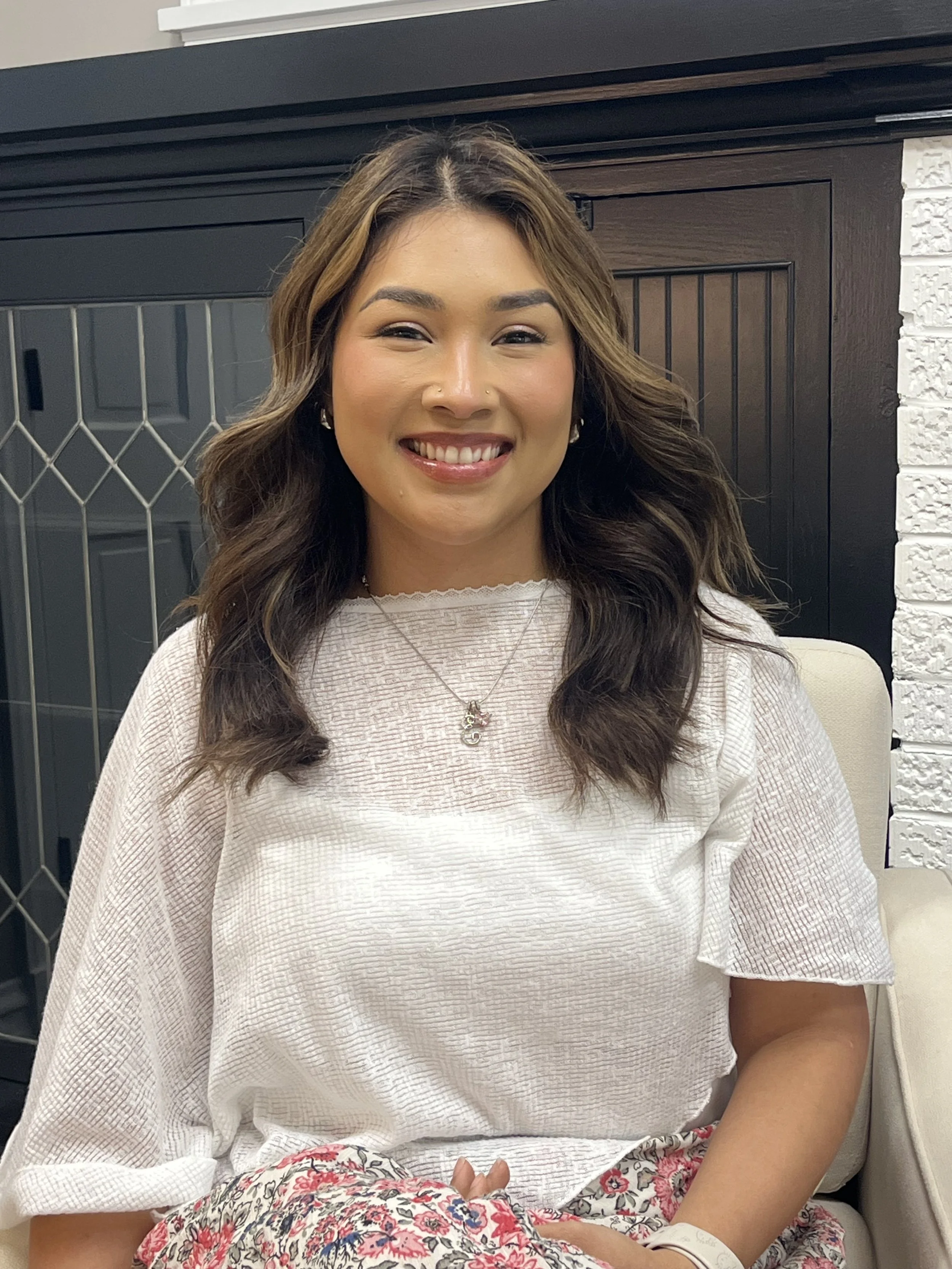 A woman with long brown hair sitting indoors, smiling at the camera, wearing a white textured top and floral pants, with a hospital bracelet on her wrist.