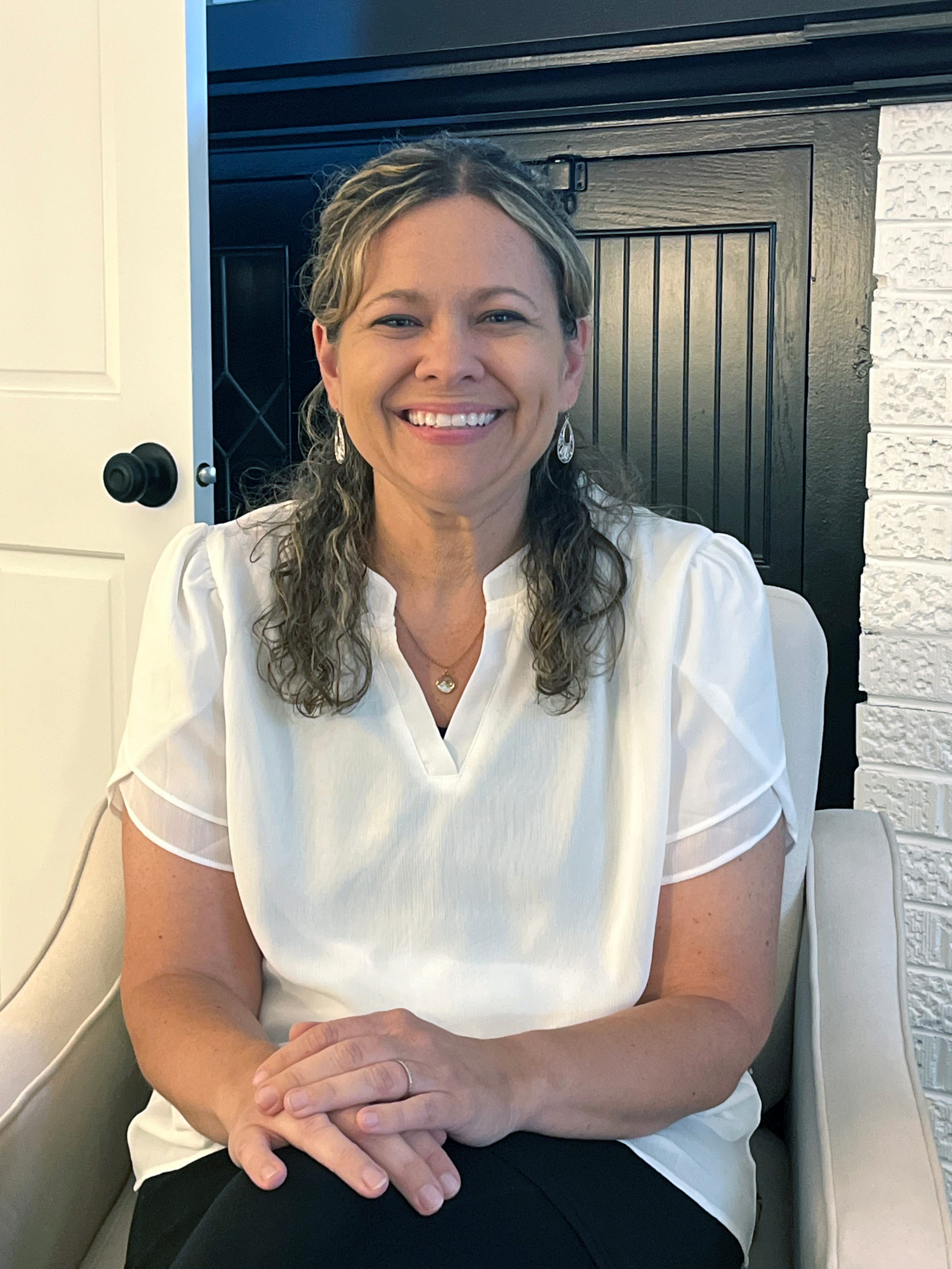 A woman with curly, blonde-tipped hair smiling, wearing earrings and a white blouse, seated on a beige chair in front of a brick and wooden wall.