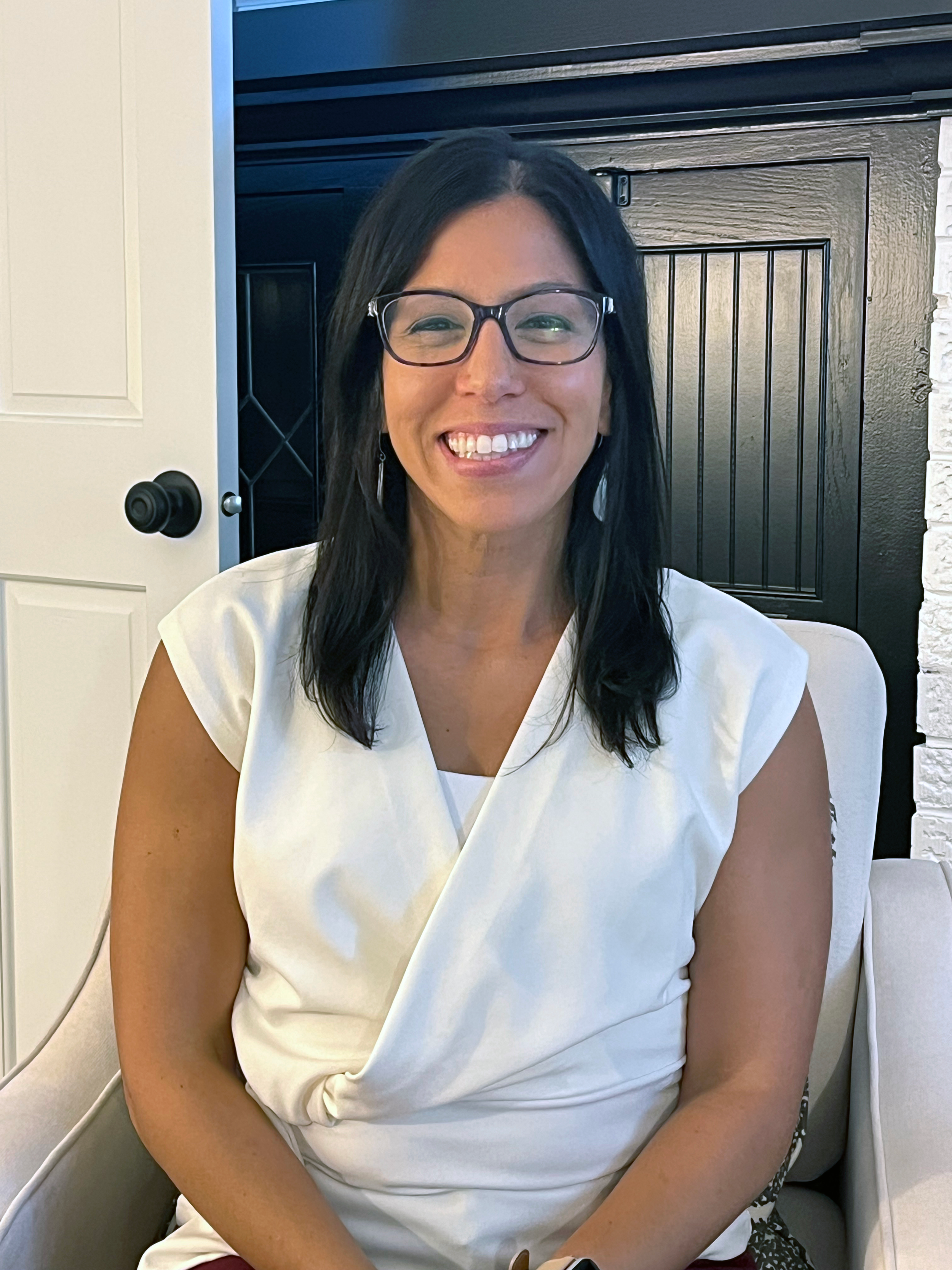 A smiling woman with dark hair, glasses, and earrings is sitting indoors in front of a black door and a white wall. She is wearing a white top.