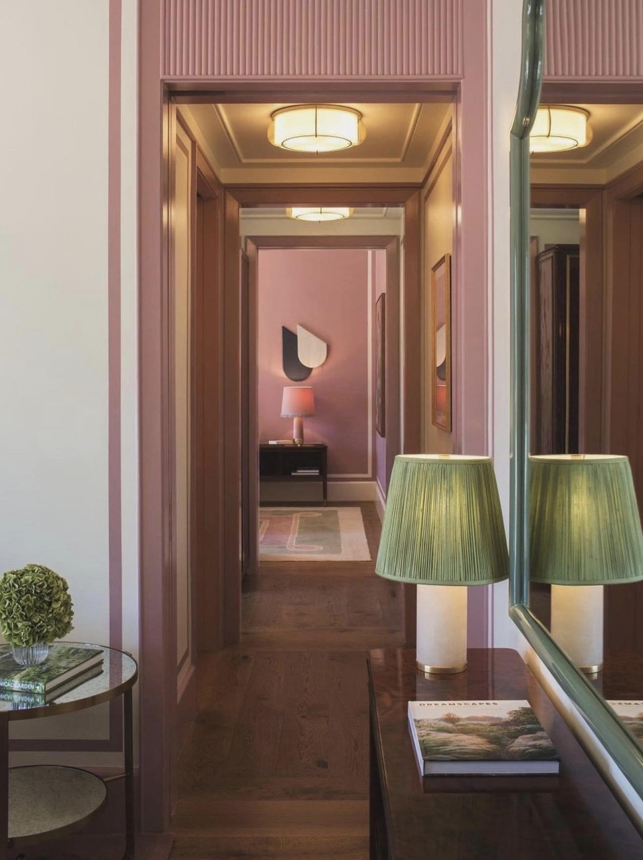 Interior view of a home with wooden floors, pink walls, and multiple ceiling lights. There is a lamp on a side table in a room at the end of a corridor, a wall art piece, and a mirror reflecting two green lamps and a shelf with books.