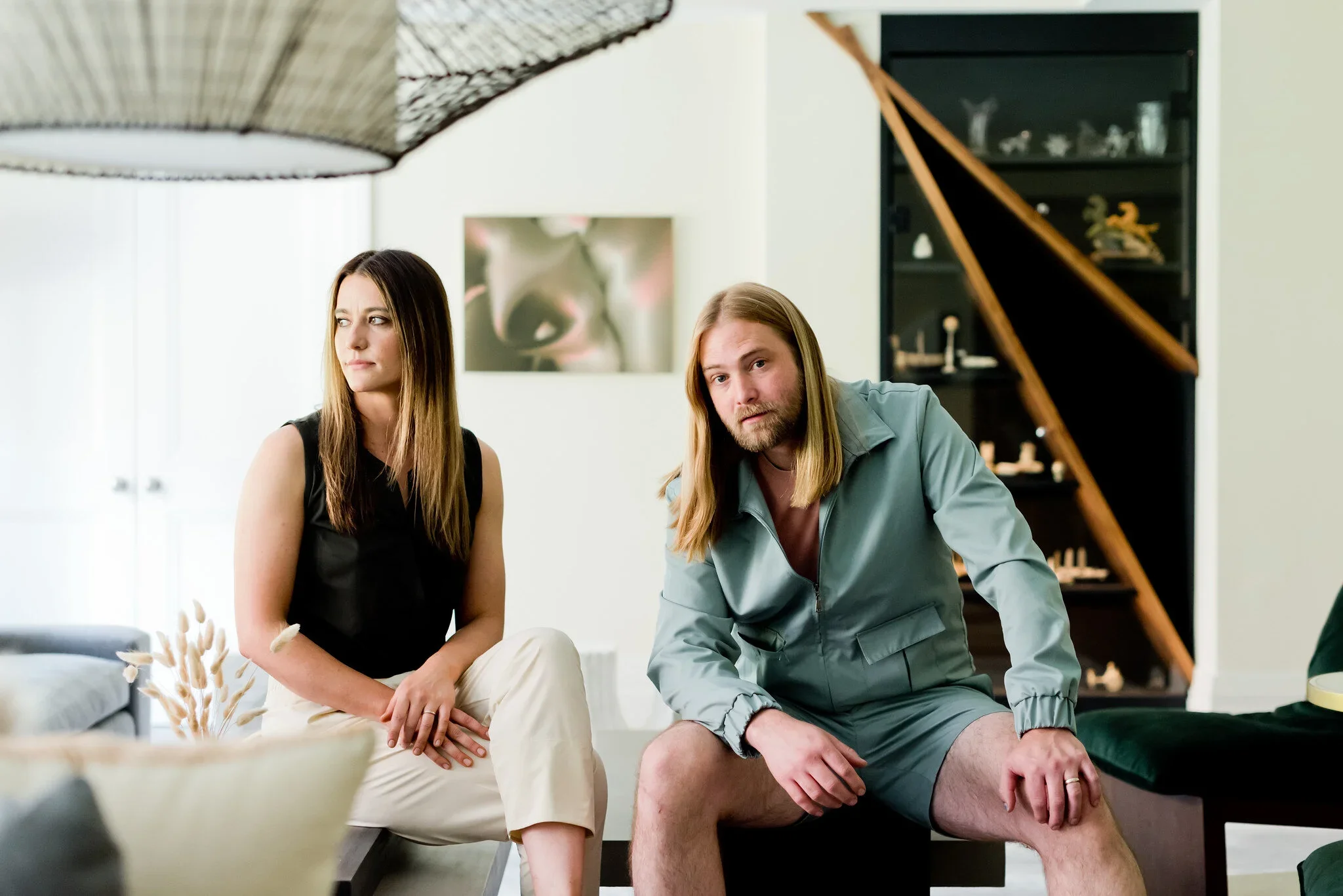 A woman and man sitting on a couch in a modern living room. The woman has long, straight hair and is wearing a black sleeveless top, looking to her left. The man has long hair, a beard, and is wearing a gray jacket and shorts, looking toward the camera. A black bookshelf with decorative items is in the background.