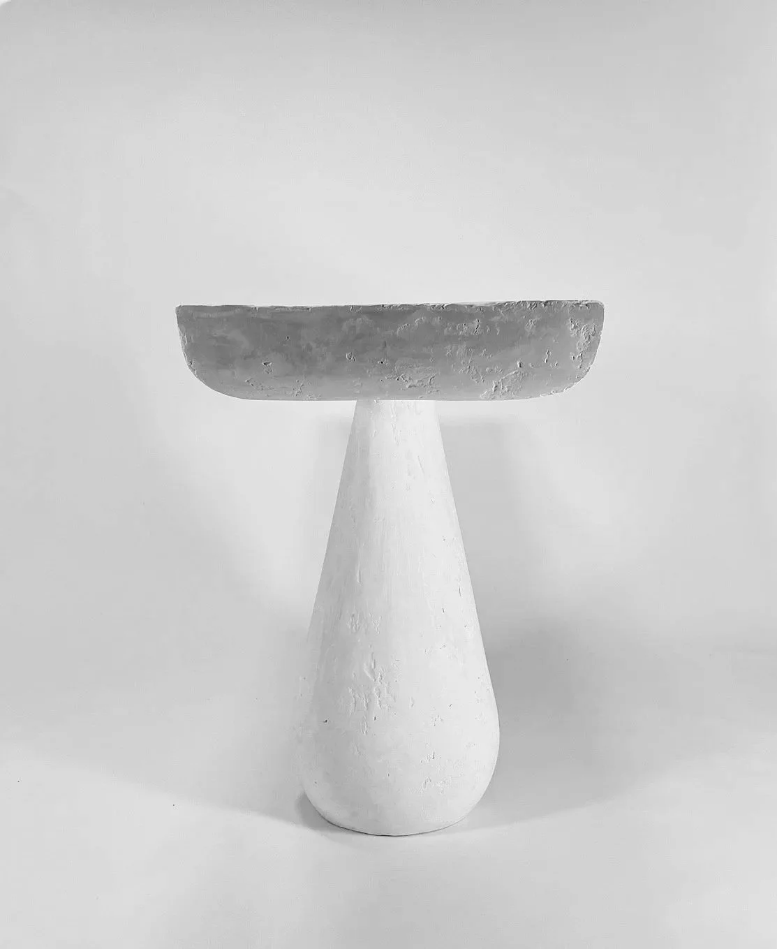 Black and white photo of a modern, minimalist pedestal with a round, shallow bowl on top. The pedestal has a tapered, smooth surface, and the background is plain and light-colored.