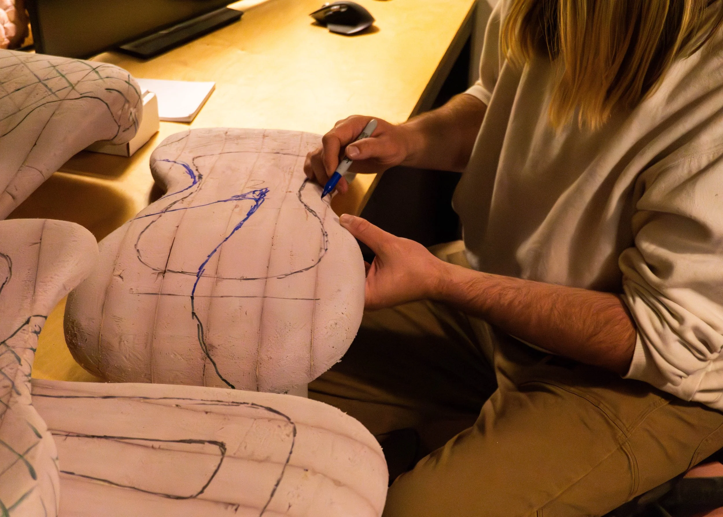 Person working on a foam mannequin torso with marked guidelines, using a pen, at a wooden table with a mouse, notebook, and computer in the background.