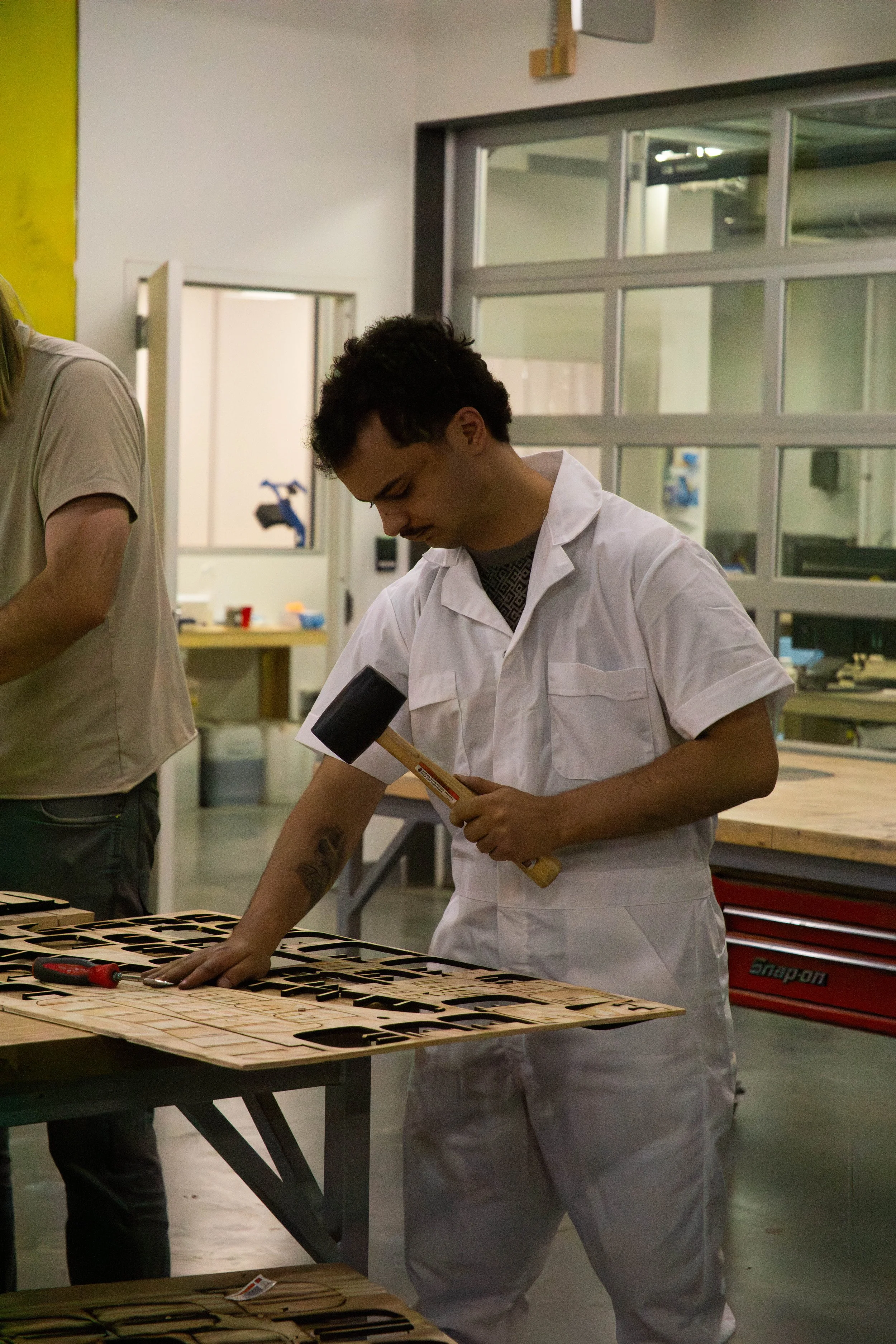 A person wearing white attire working with wooden pieces on a table in a workshop or lab environment, with tools and shelves in the background.
