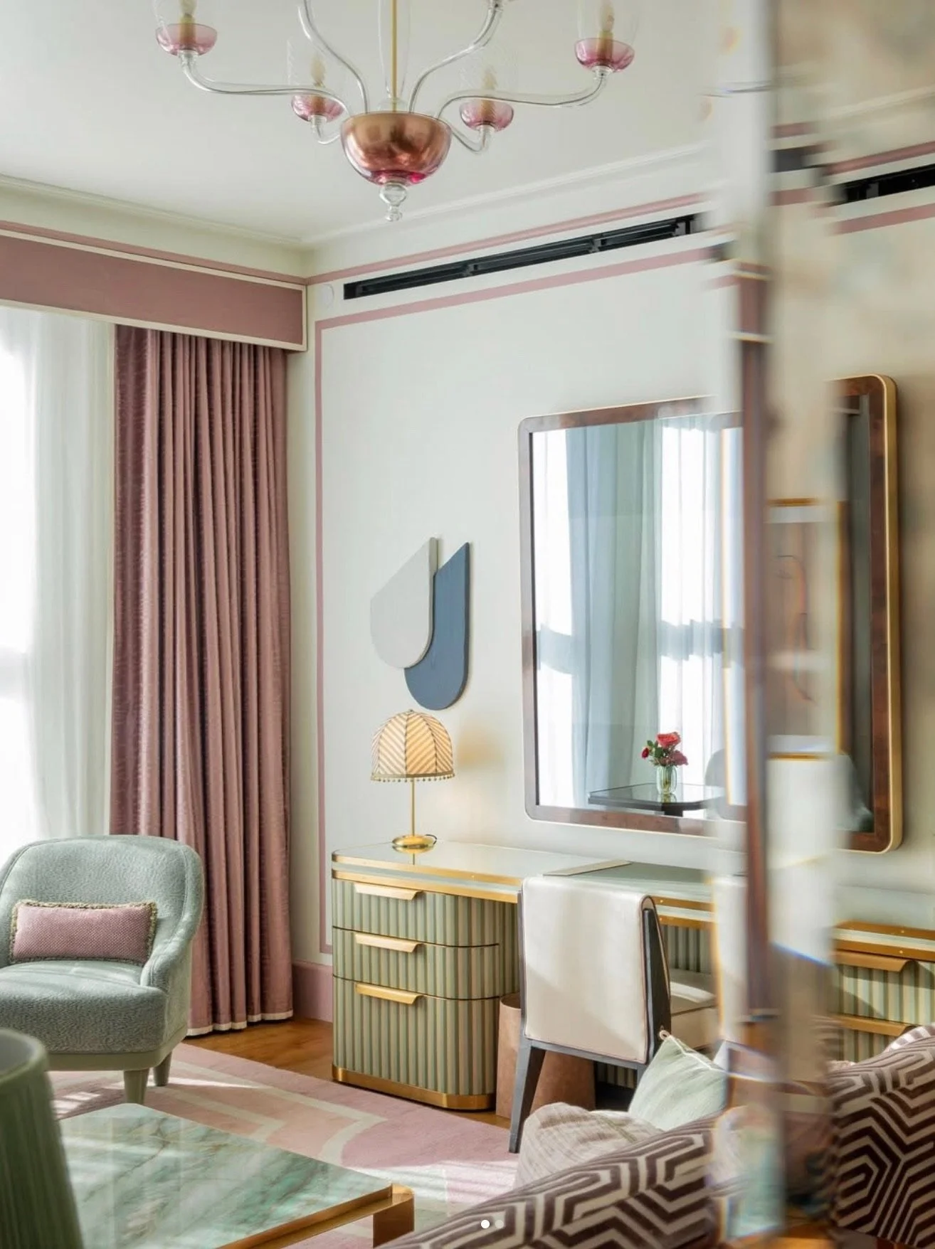 Living room with pastel pink, green, and white decor, featuring a pink curtain, a green armchair, a vintage beige and pink striped dresser, a mirror with a wooden frame, modern lamps, and a small table with flowers.
