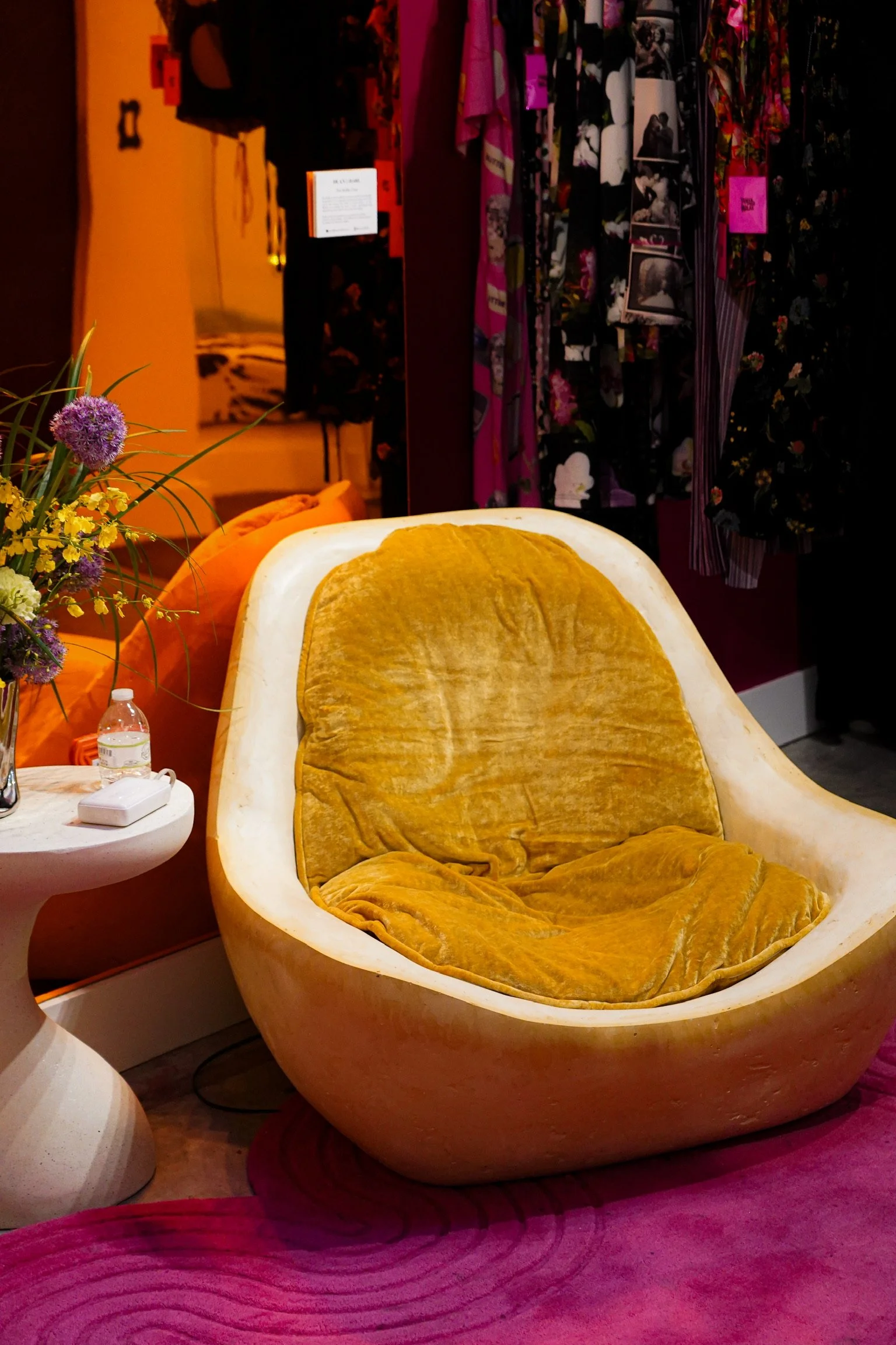 A yellowish-orange velvet chair resembling an egg shape with a rounded back and inside-cushion, placed on a pink circular rug with decorative circular patterns. To the left, a side table with a floral arrangement and a water bottle. In the background, a clothing rack with various colorful garments, and a partially visible bedroom with yellow bedding, a mirror, and wall decor.