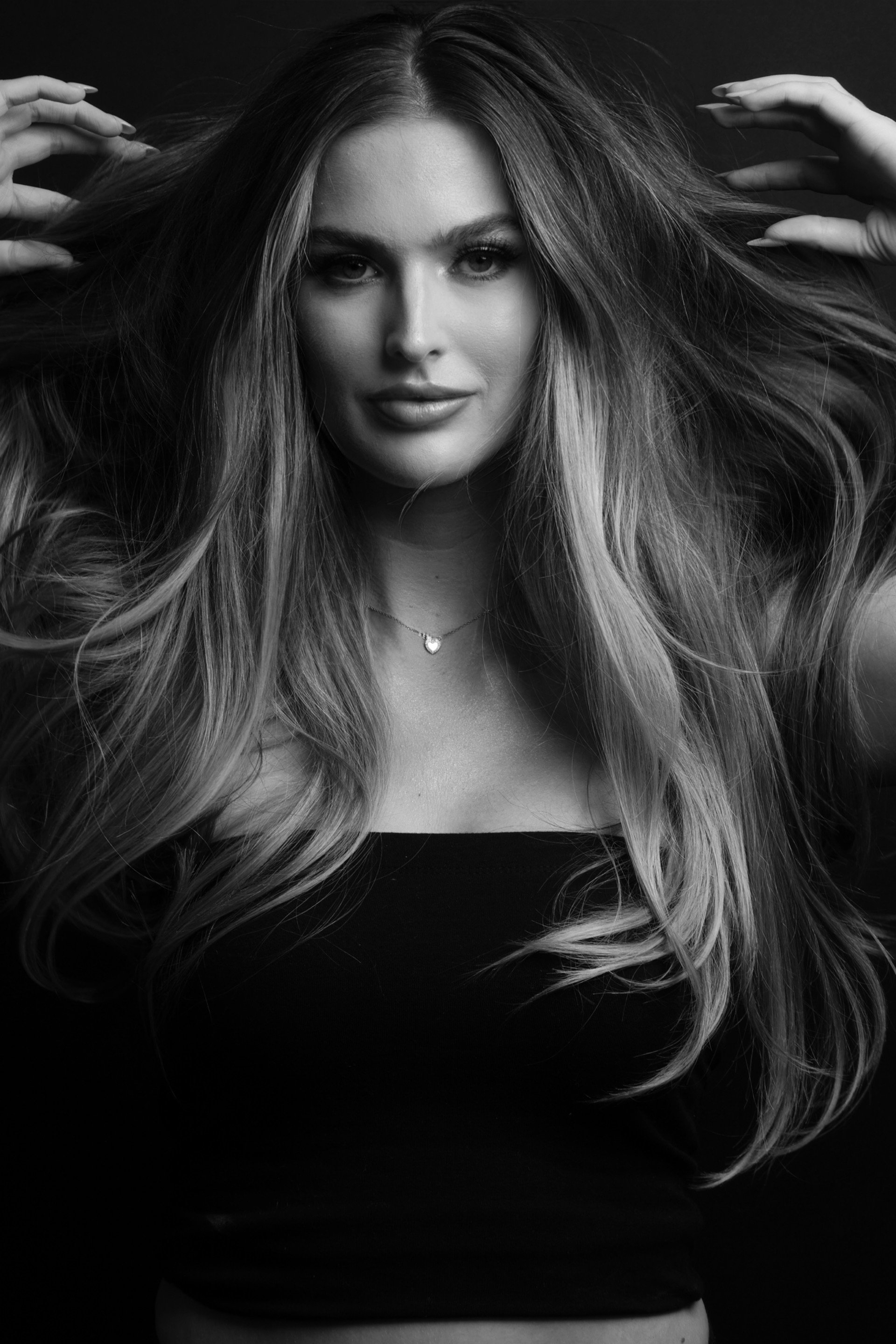 A woman with long wavy hair, looking at the camera, with her hands raised near her head, in a black and white studio portrait.