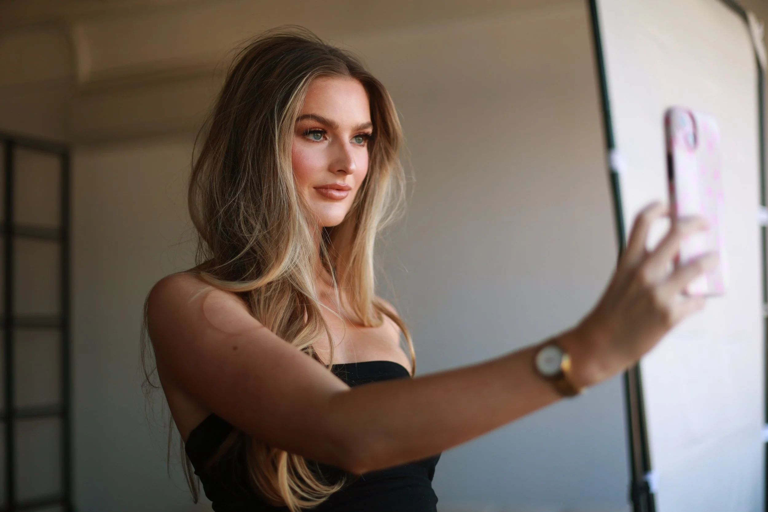 A young woman with long, wavy blonde hair takes a selfie in a mirror, wearing a black strapless top, with a neutral background and a blurred phone in her hand.