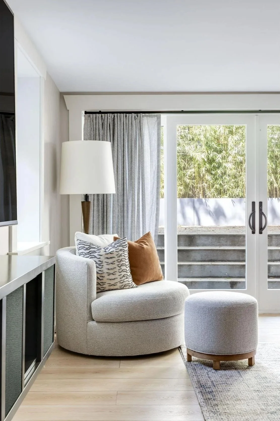 Living room renovation with a modern armchair and matching ottoman near glass sliding doors, gray curtains, a white floor lamp, and outdoor greenery visible through the doors.