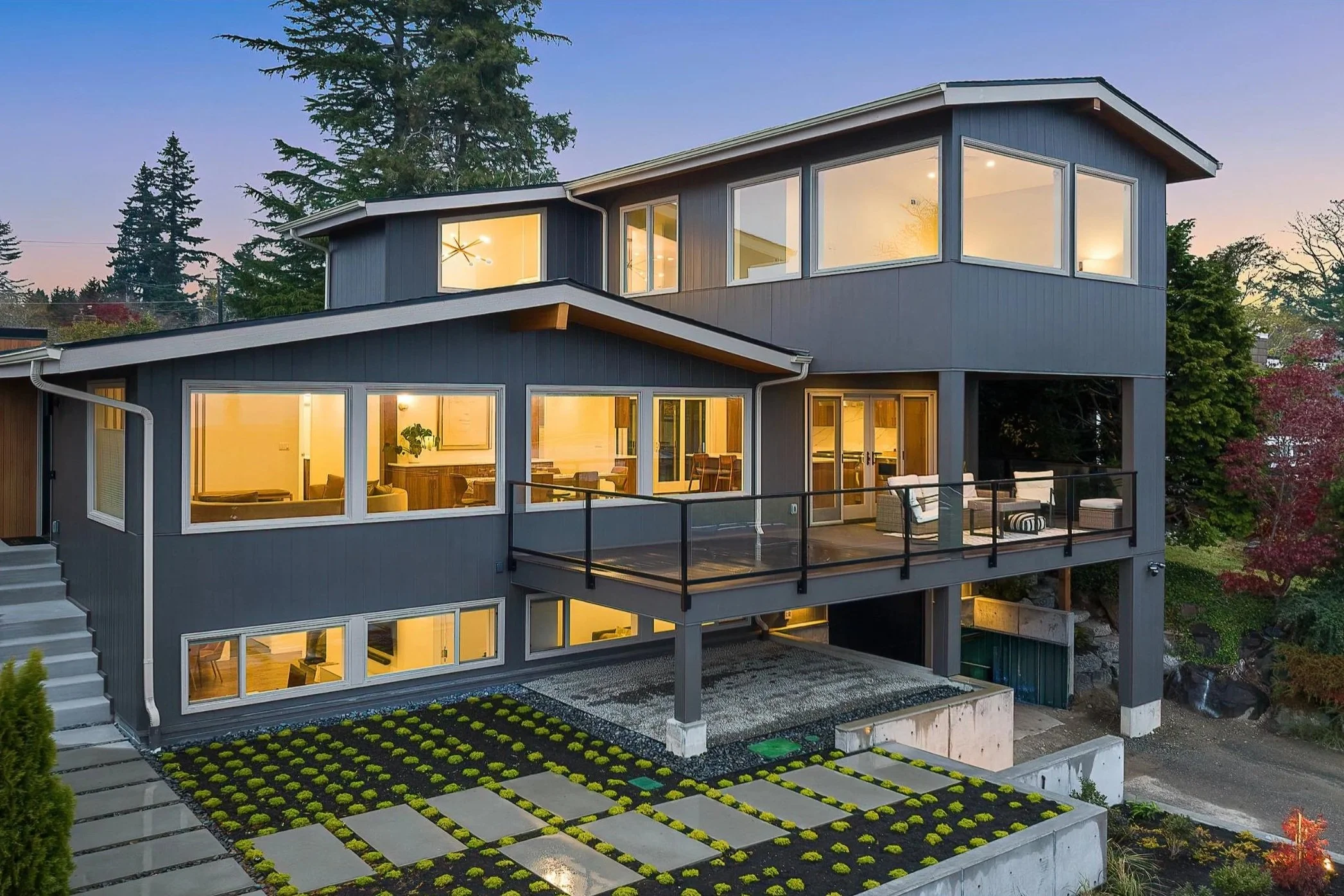 Multi-story modern house addition and renovation with large windows, illuminated interior, surrounded by trees and a small driveway with a patterned garden in the foreground.