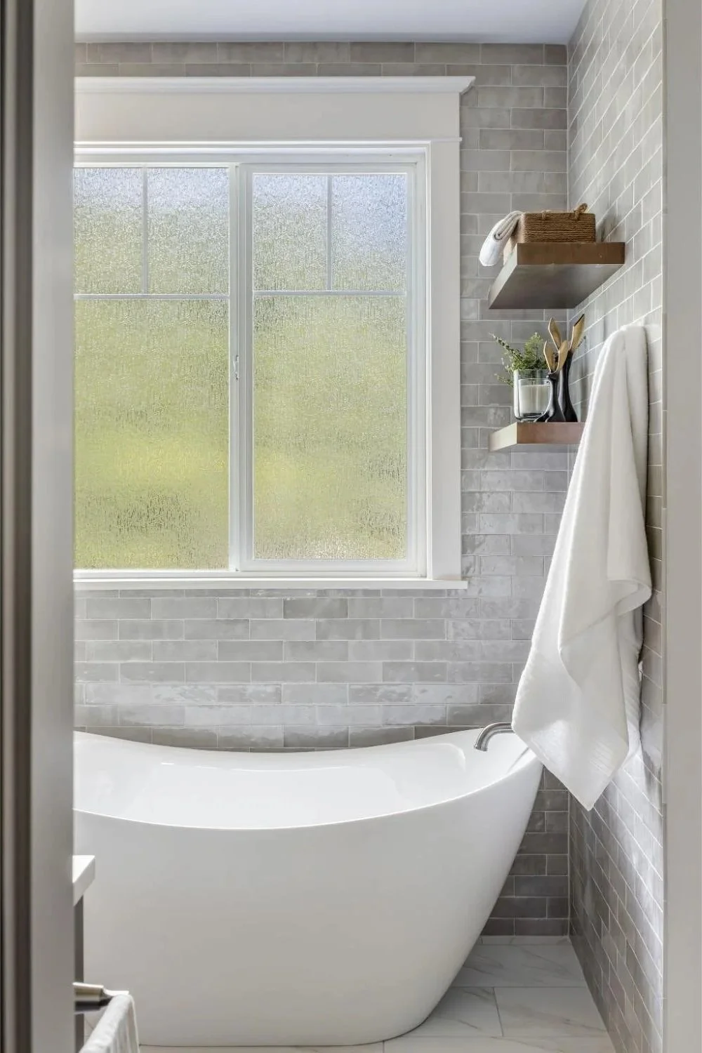 A modern bathroom renovation with a freestanding bathtub, a large frosted window, and floating shelves with decor and a towel.