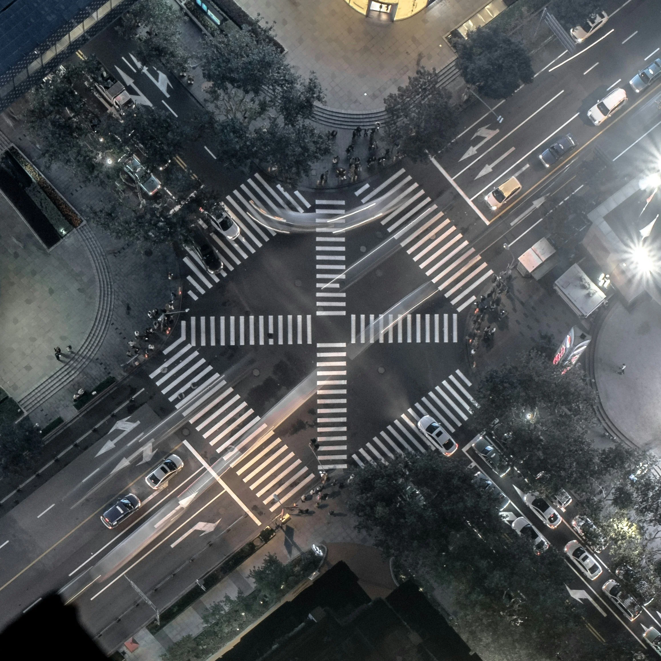 An aerial view of a city street at night showing a crosswalk, parked cars, trees, and people walking on the sidewalk near a building.