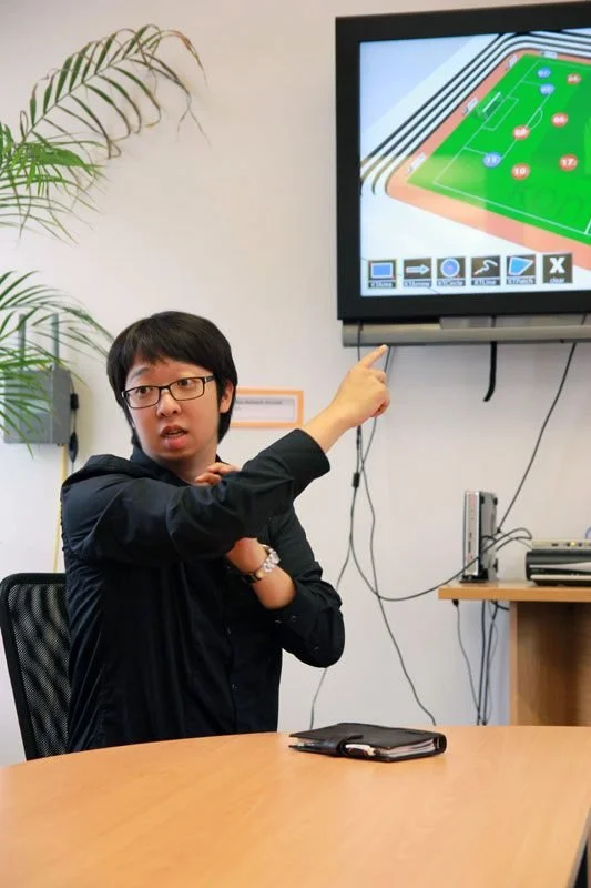 A person with glasses and a black jacket sitting at a table, pointing at a television screen displaying a diagram of a soccer field with player positions.