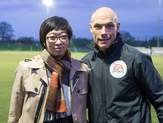 Two men standing outdoors on a sports field during dusk, with bright stadium lights in the background. One man is wearing glasses, a beige trench coat, and a multicolored scarf. The other man is wearing a black EA Sports jacket and is bald.