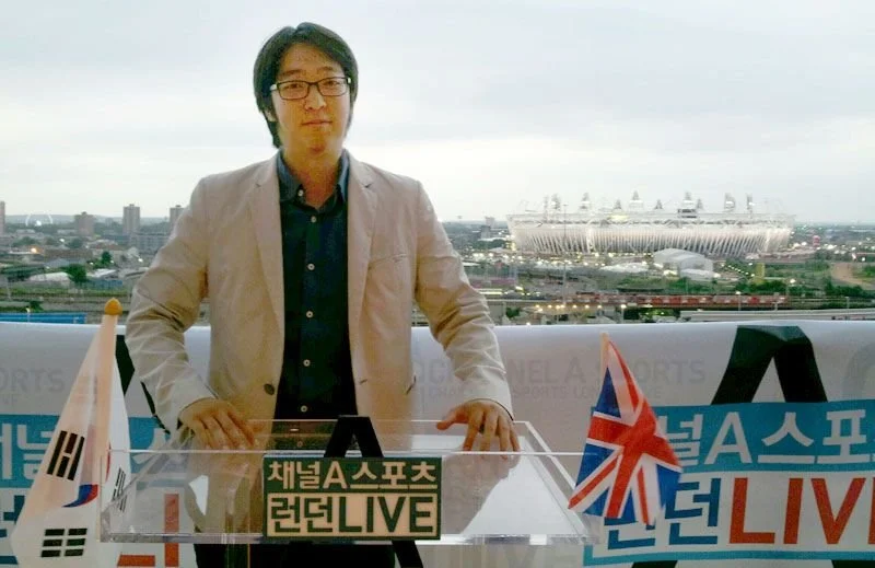 The company's founder wearing glasses and a beige blazer standing in front of a sports broadcast setup, with South Korea and United Kingdom flags on either side, and a cityscape with a large London Olympic Stadium in the background during daytime.