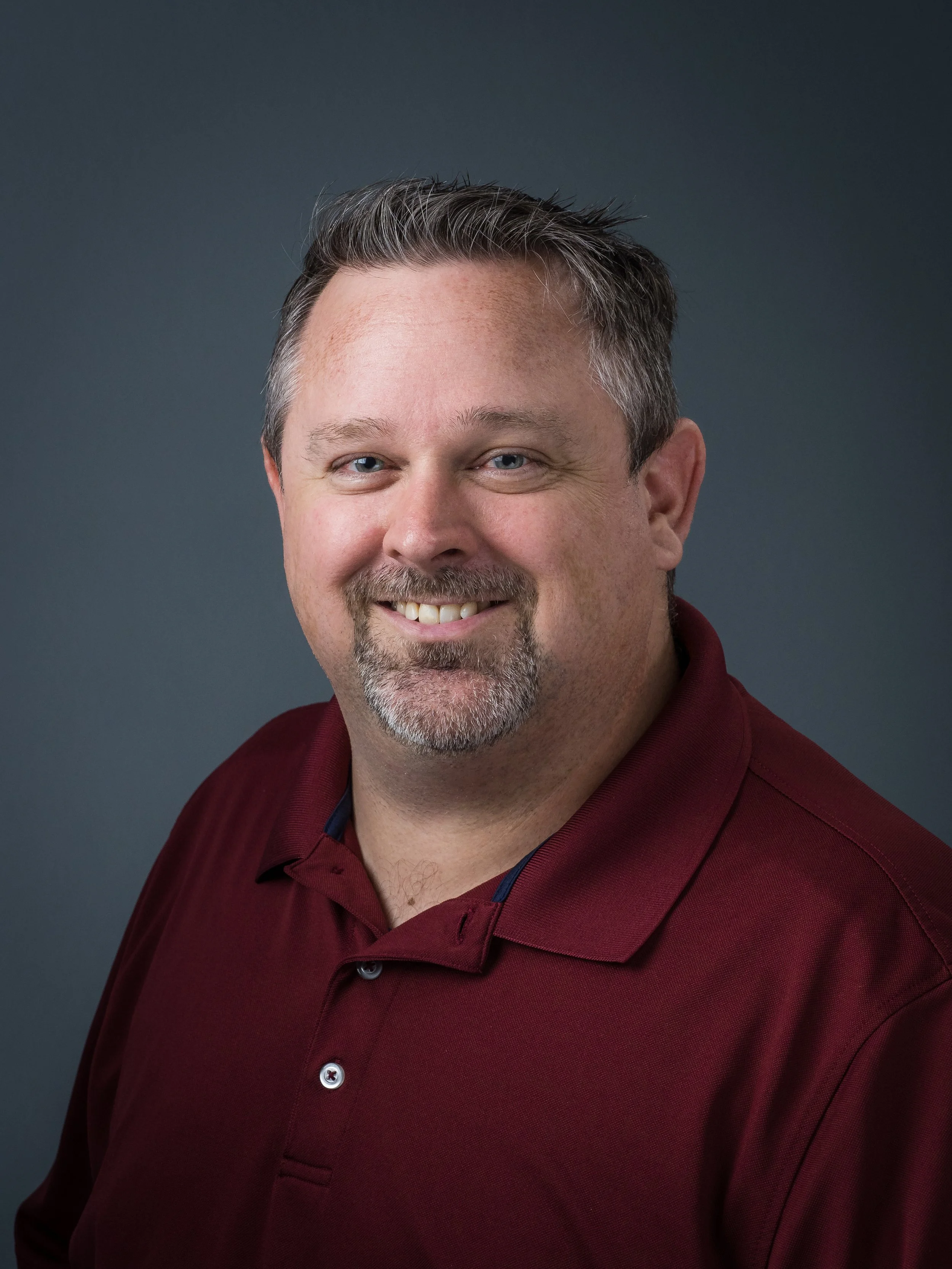 Close-up of a smiling man with short, graying hair, beard, and blue eyes, wearing a maroon polo shirt against a dark gray background.