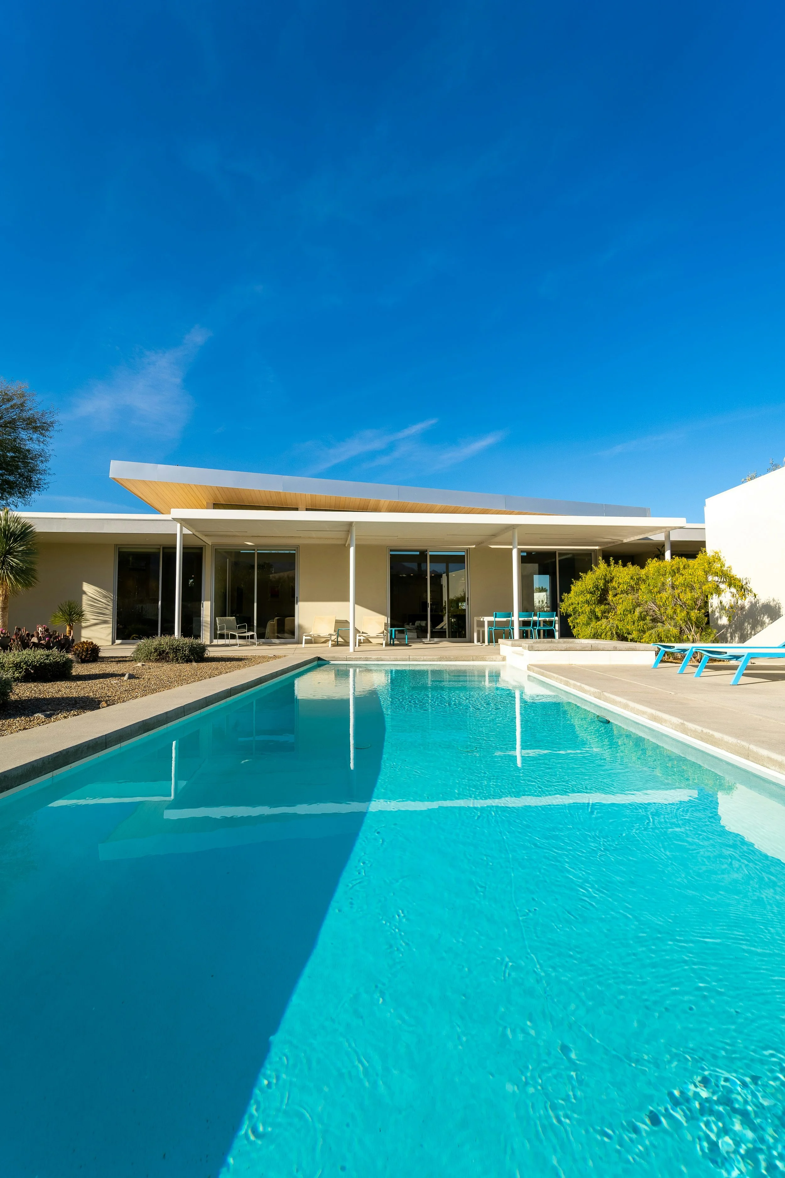 Modern house with a swimming pool in the backyard, surrounded by chairs and landscaping, under a clear blue sky.