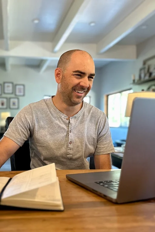 A smiling man, wearing a light gray t-shirt, sitting at a wooden table in a bright room, looking at a laptop. There is an open book in front of him, and the room has a high ceiling with exposed beams and framed pictures on the wall.