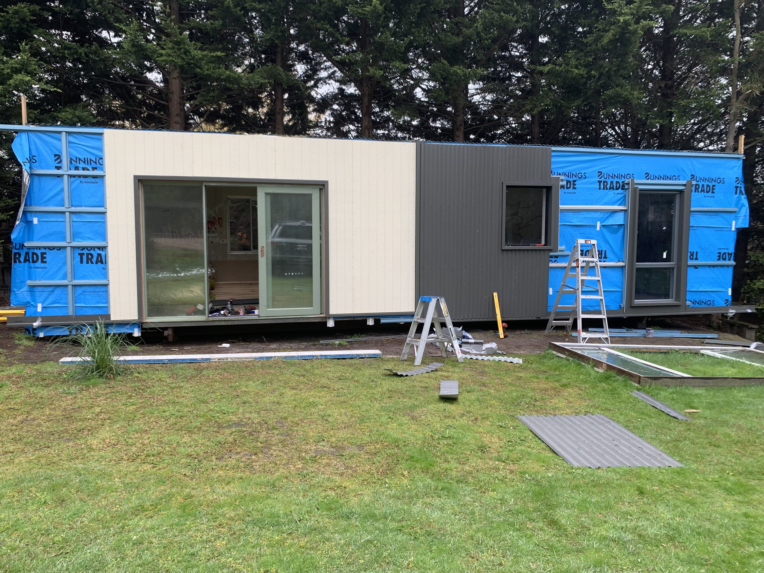 A house under construction with partially installed exterior siding, window, and construction tools and materials outside on the grass with trees in the background.