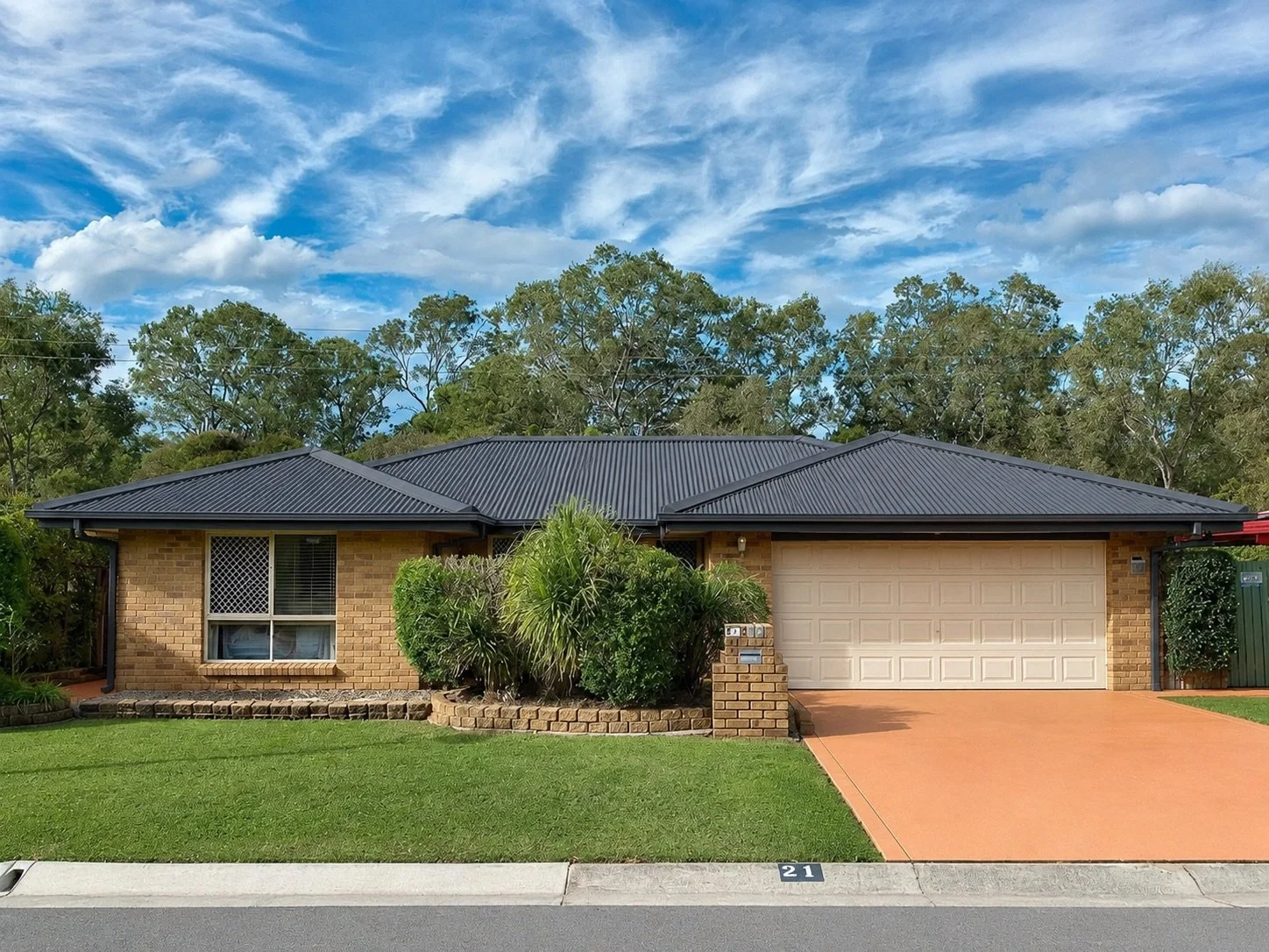 Single-story brick house with a black metal roof, white garage door, and landscaped front yard with green grass and bushes, in a suburban neighborhood under a partly cloudy sky.