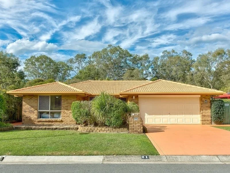 Single-story brick house with a tiled roof, front yard with grass and bushes, driveway leading to a garage, under a blue sky with clouds.