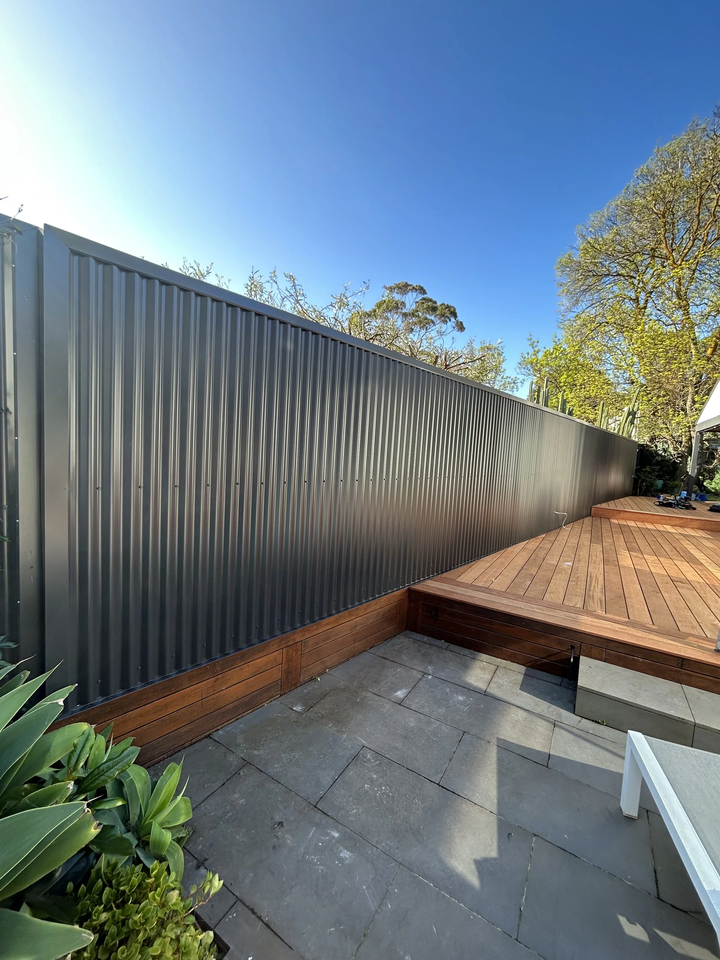 A backyard patio with a wooden deck and a tall metal fence, surrounded by plants and trees under a clear blue sky.