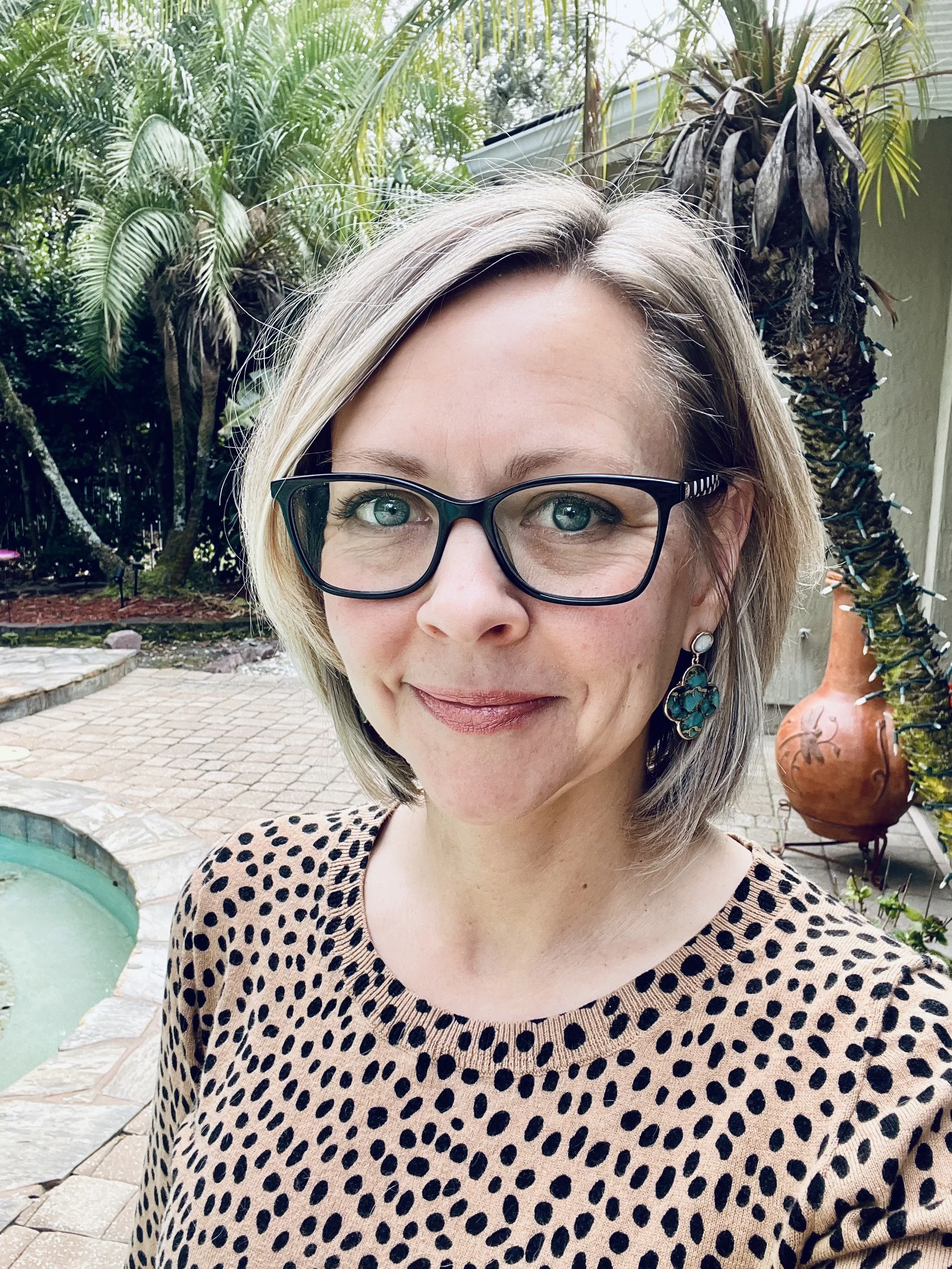 Close-up of a woman wearing black-rimmed glasses and patterned top, standing outdoors with lush green plants and a clay pot in the background.