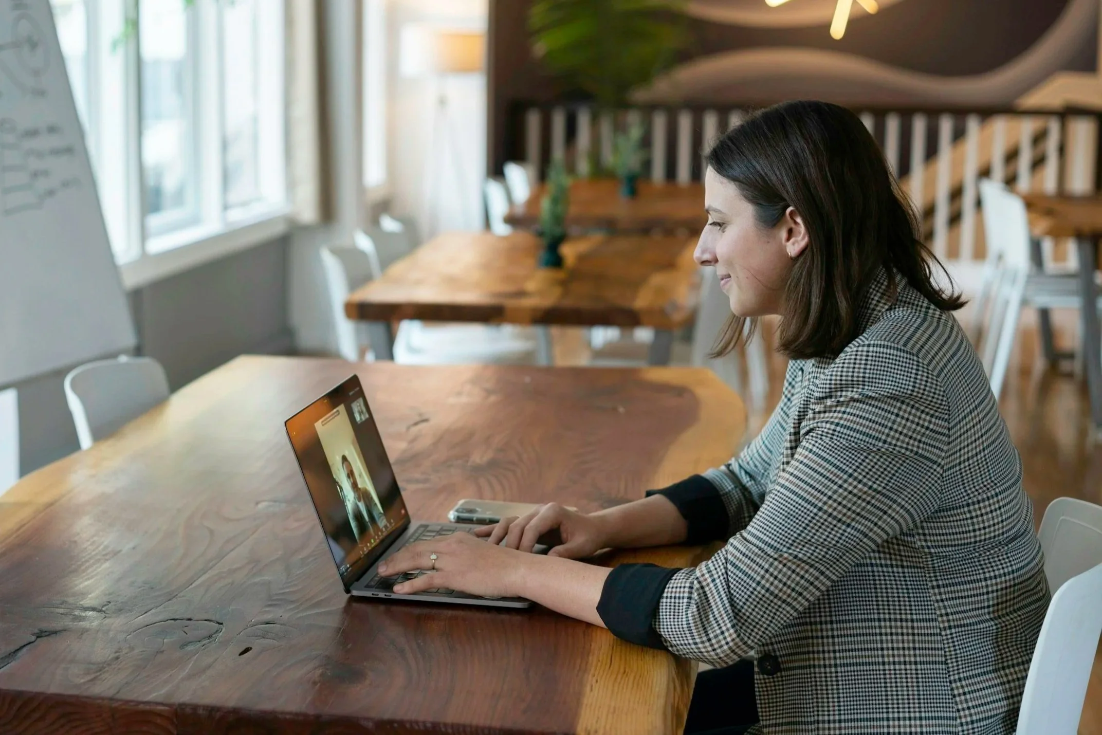 A woman with dark hair sitting at a large wooden table, smiling and looking at a laptop screen. The room has natural light coming through the windows, with white chairs and a whiteboard in the background.