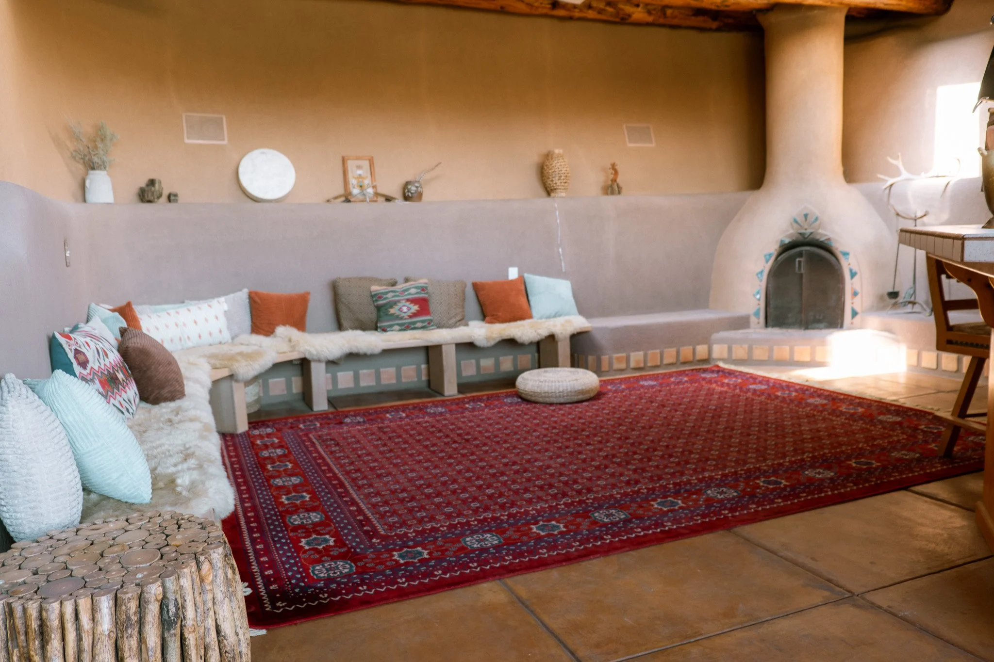 Living room with a red patterned rug, a long bench with colorful pillows, a round cushion, a wood stump side table, and decorative items on the wall shelf including vases, a picture frame, and small sculptures. There's a built-in fireplace with a unique curved shape and turquoise tile accents, and a window with light coming through.