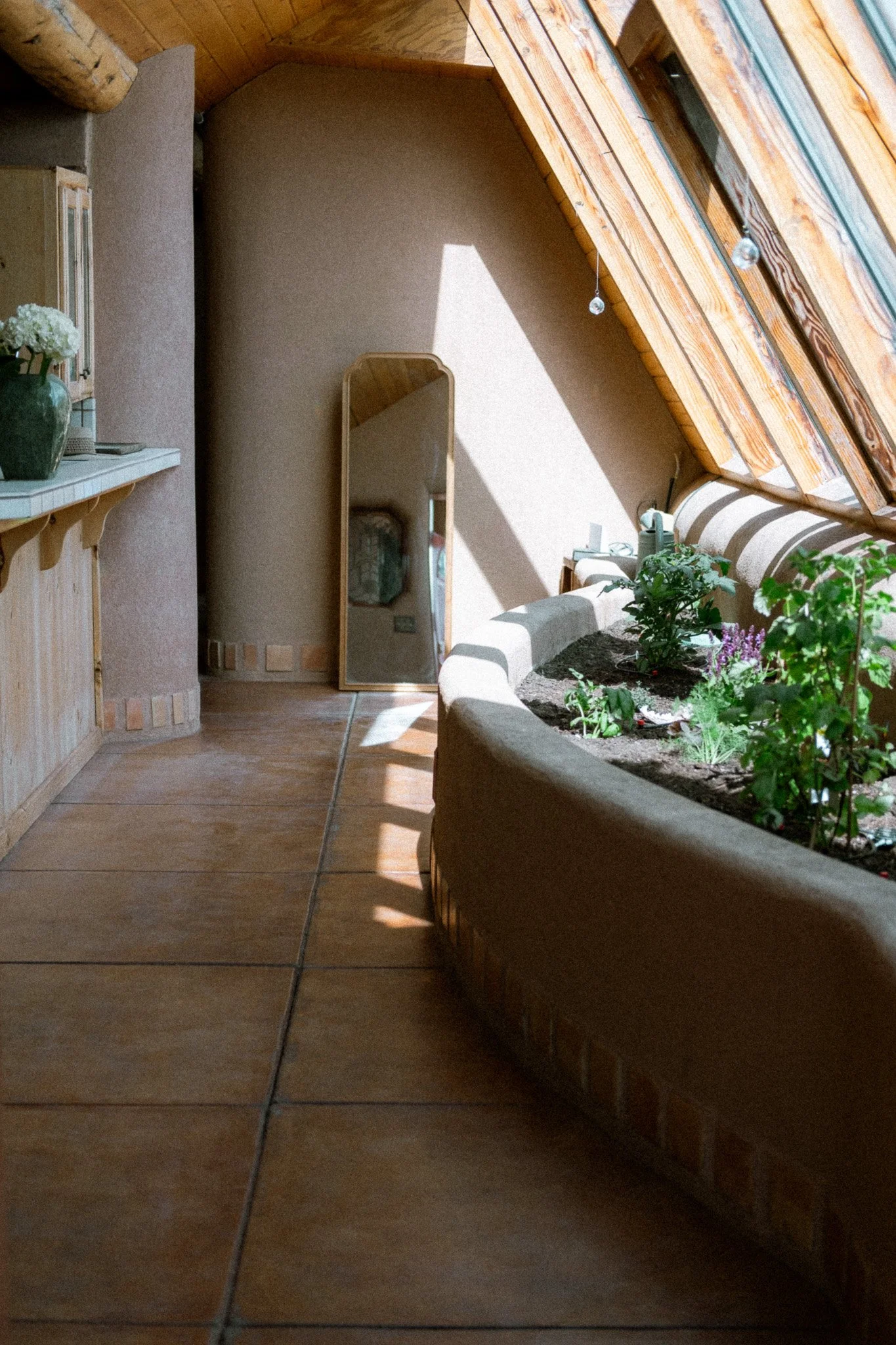 Interior of a room with a sloped wooden ceiling and a skylight window. Contains potted plants, a mirror, a bookshelf with a vase of flowers, and a cozy sofa.