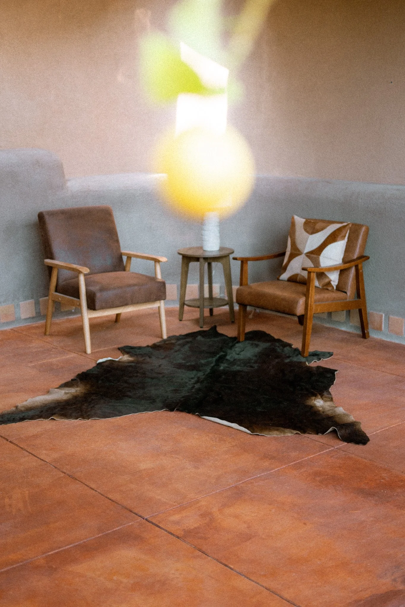 Living room with two wooden armchairs with cushions, a small side table with a lamp, a black and white animal hide rug on a terracotta tile floor, and a wall decorated with a simple neutral color.