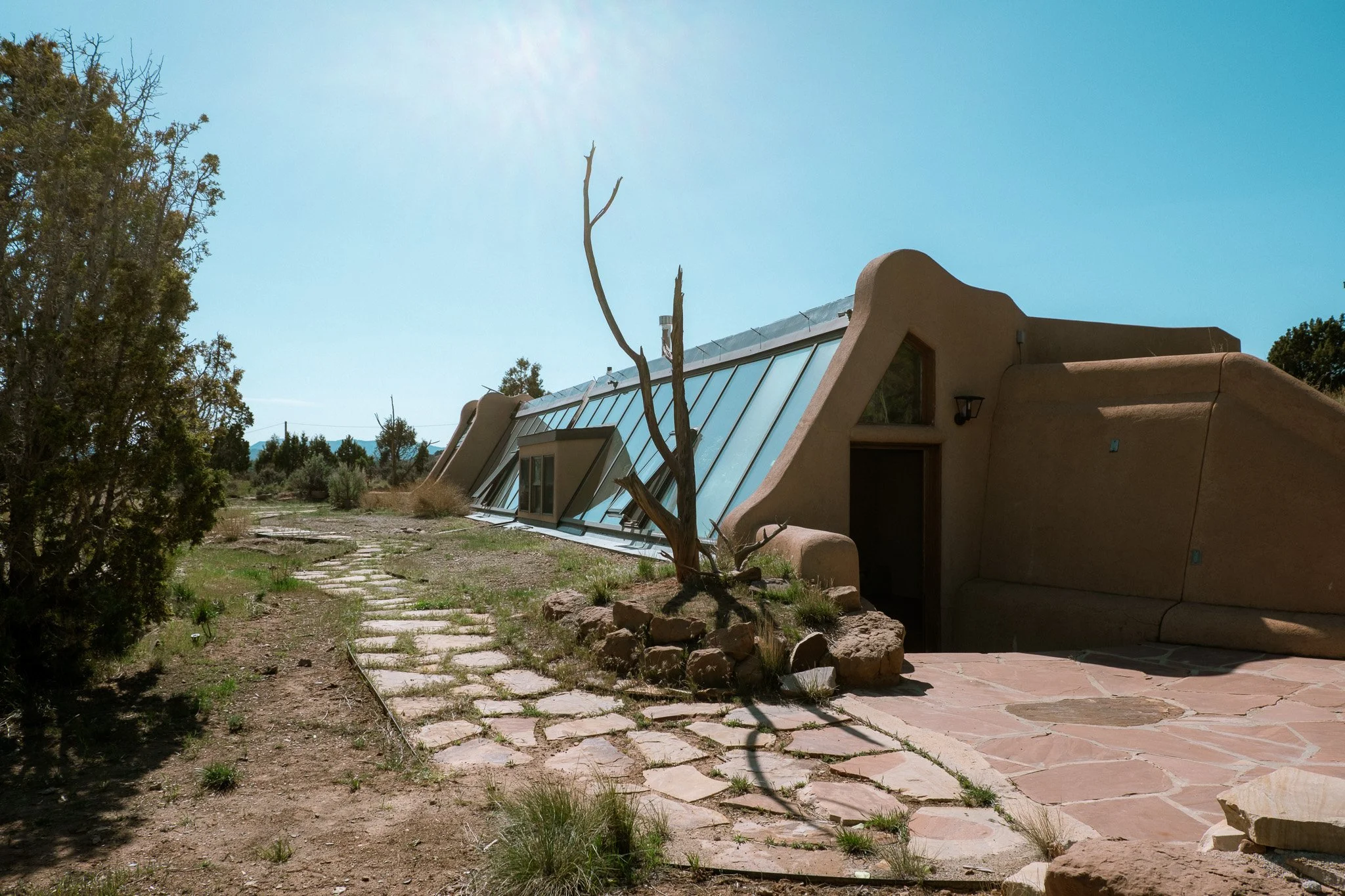 A desert scene with a modern, adobe-style house featuring a sloped roof with large glass windows, surrounded by sparse vegetation and a dirt pathway with stepping stones. A leafless tree stands in front of the house under a bright, clear sky.
