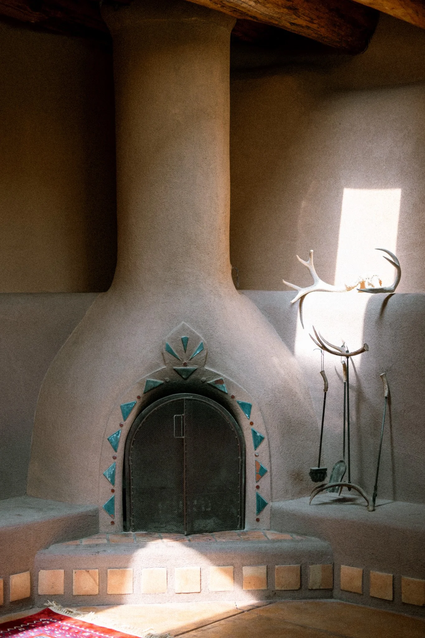 A corner fireplace with a rounded top, decorated with turquoise and orange tiles around the arched metal fire door, and a pair of antlers mounted on the wall to the right, with sunlight casting a rectangular light pattern on the wall.