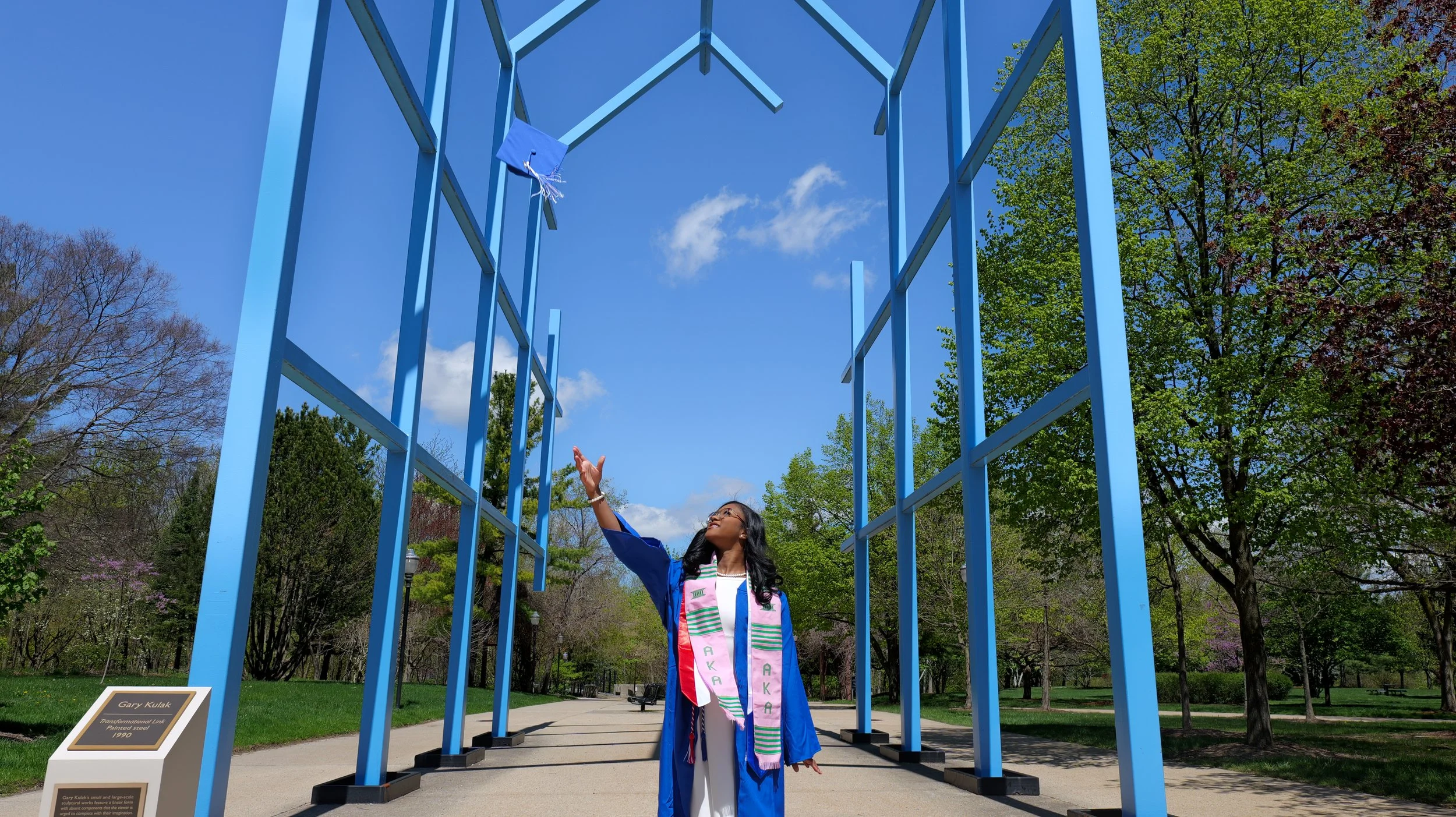 A young woman in graduation cap and gown is standing outdoors under a tall blue structure, reaching up towards the sky with one hand, celebrating her graduation with trees and blue sky in the background.