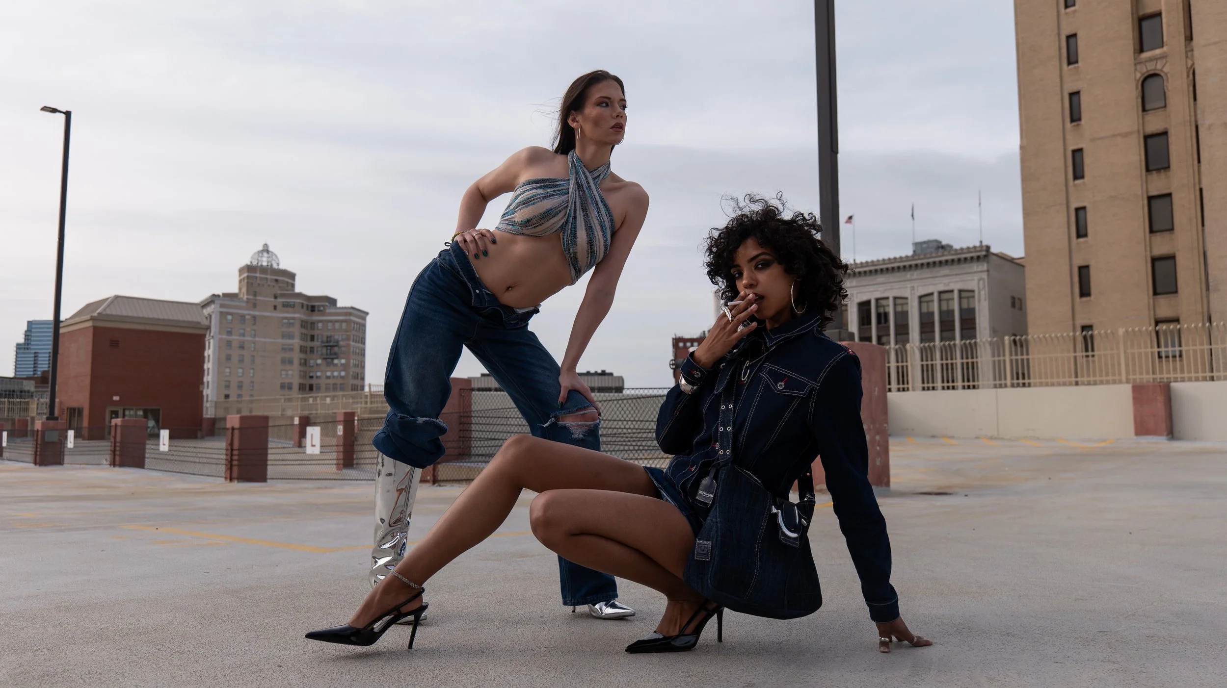 Two women posing on top of a parking garage with a city skyline background. One woman is sitting on the ground with one knee up, wearing a dark denim jacket, shorts, and high heels, smoking a cigarette. The other woman is standing, leaning over with 