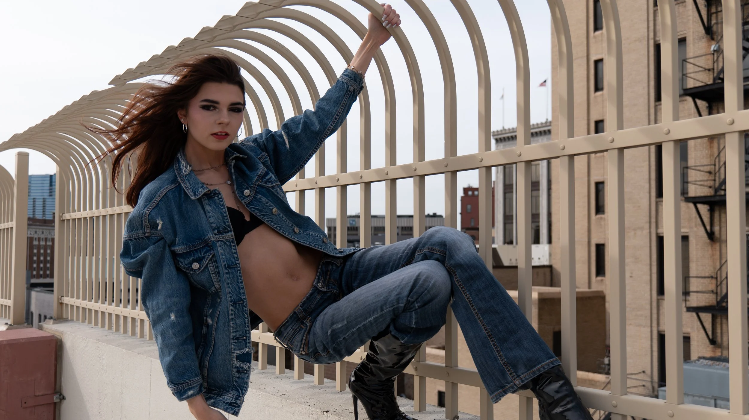 A woman wearing a denim jacket and pants sits on a rooftop with a city skyline and brick buildings in the background, holding onto a curved fence.