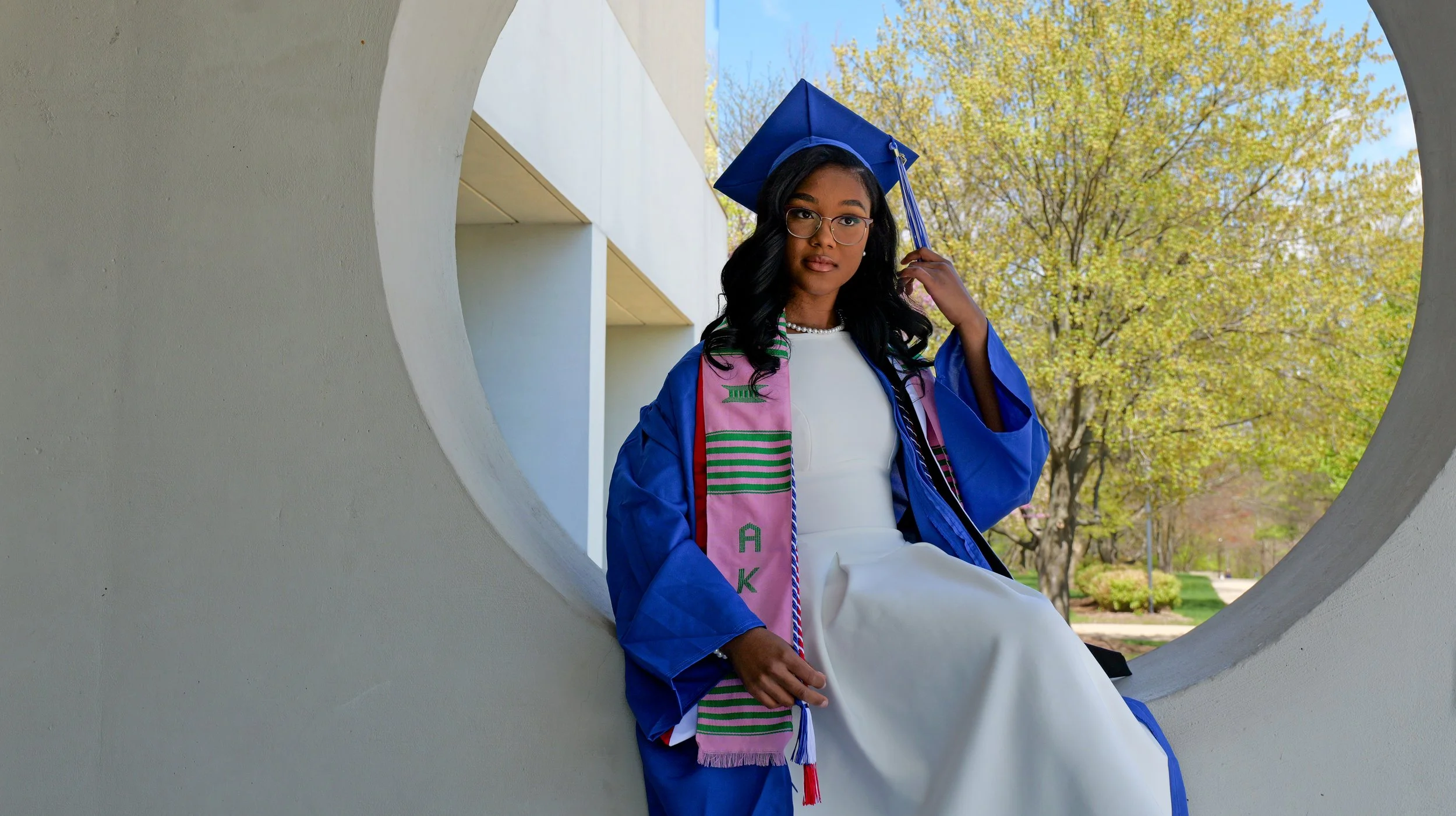 Young woman in a blue graduation gown and cap sitting on a circular concrete structure outdoors, adjusting her cap with one hand, with trees and a building in the background.
