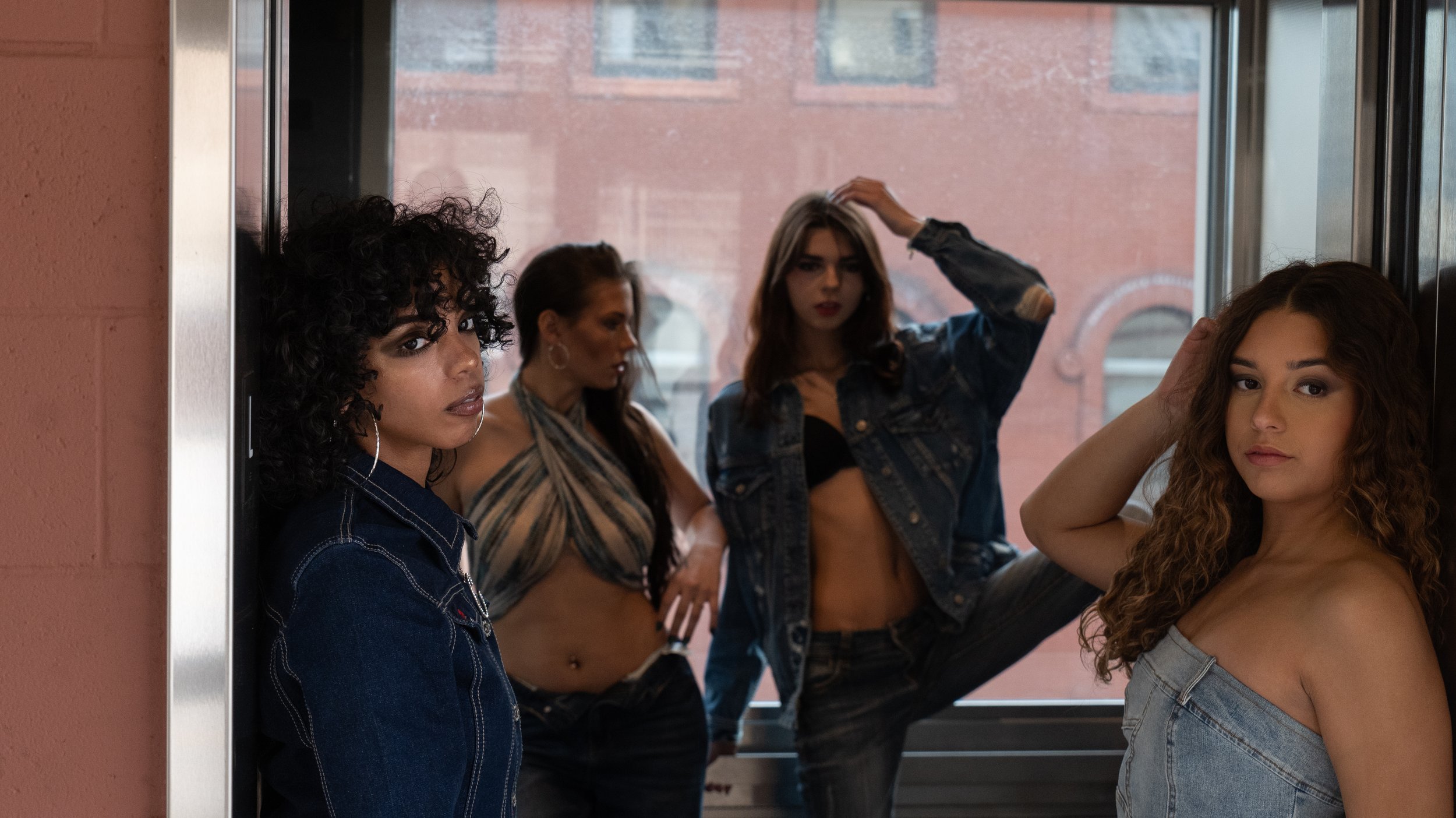 Five young women posing near an elevator, some leaning against the wall or sitting, with a brick building visible outside the window. They have various hairstyles and casual clothing.