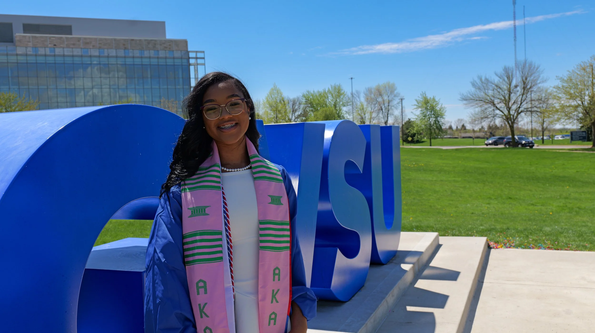 A young woman wearing graduation attire and a pink stole with green lettering stands outdoors in front of large blue letters with a city building and trees in the background.