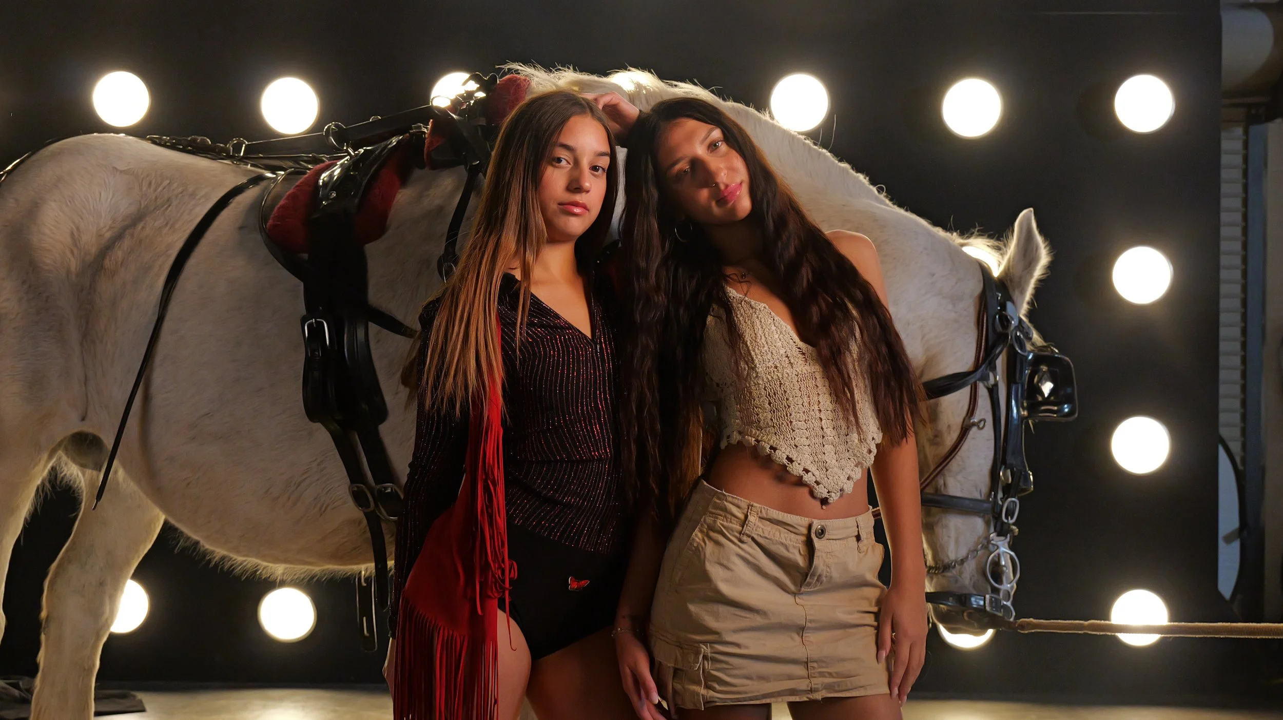 Two young women posing in front of a white horse with a harness, in a setting with bright circular lights behind them.