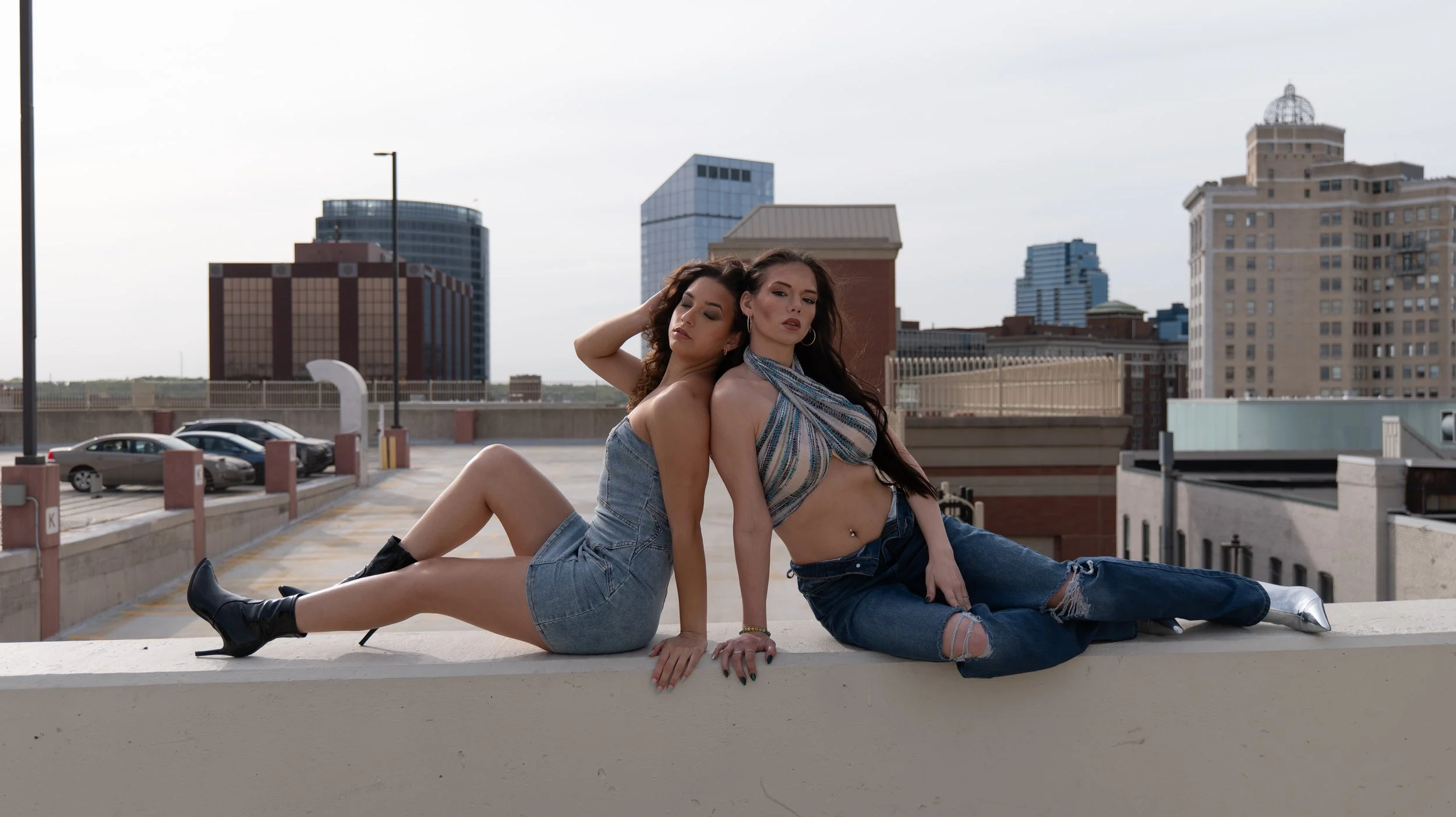 Two women sit on a rooftop ledge with an urban cityscape in the background, dressed in denim outfits and boots, posing for a photo.