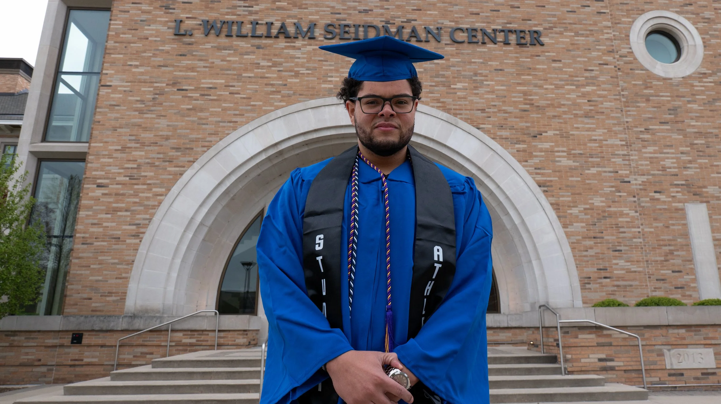 Graduate in blue gown and cap standing in front of L. William Seidman Center