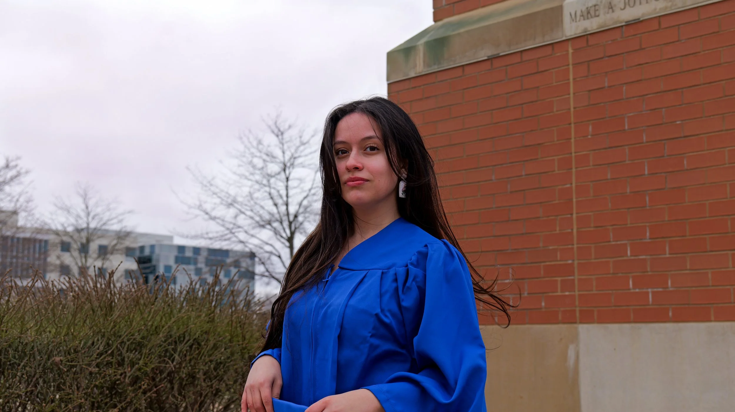 Young woman with long dark hair wearing a blue graduation gown standing outdoors by a brick building with leafless trees and city buildings in the background.