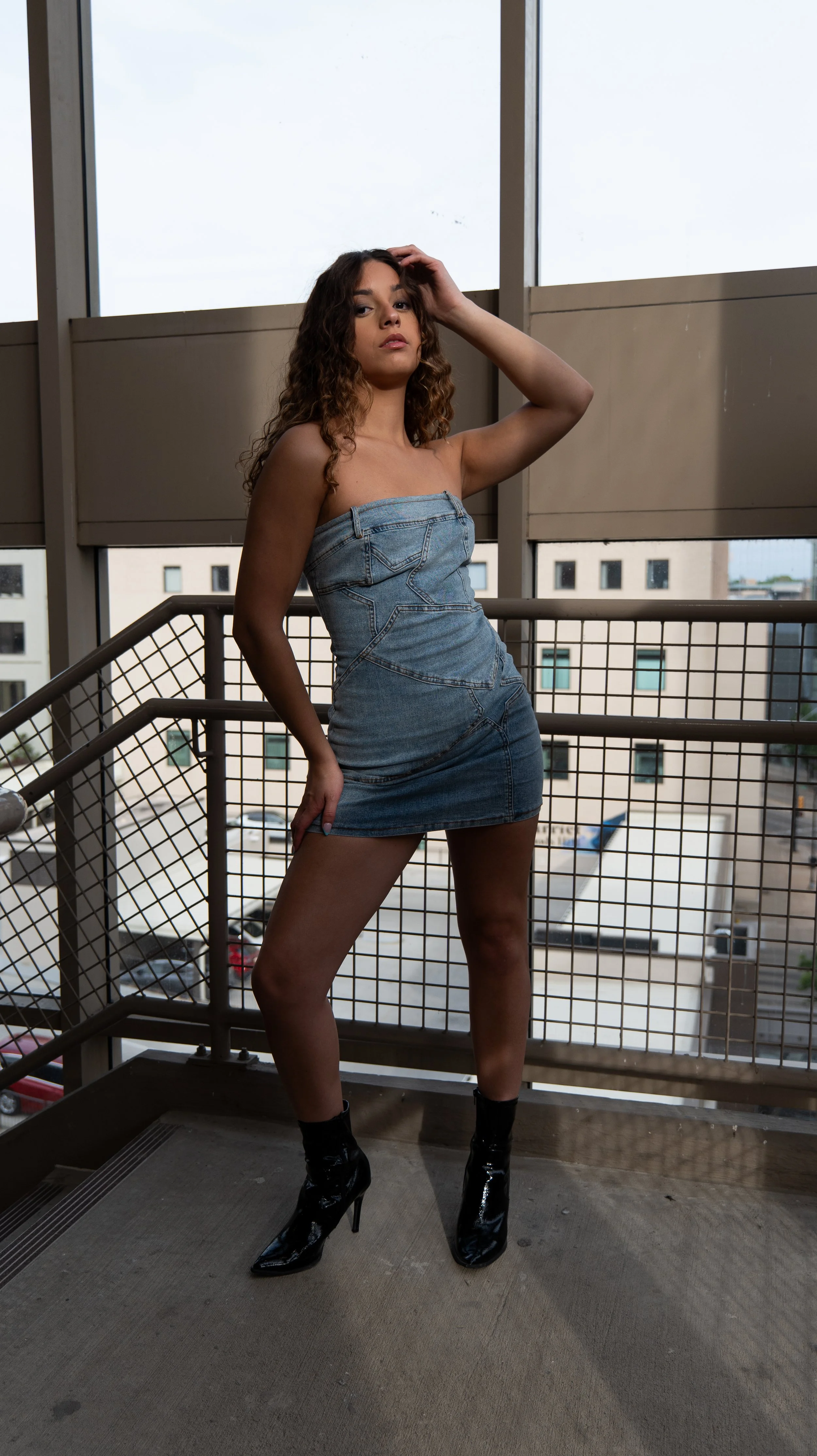 A woman with curly brown hair in a strapless denim mini dress and black high-heeled boots standing on a balcony with a metal railing, overlooking a parking lot and buildings with windows.