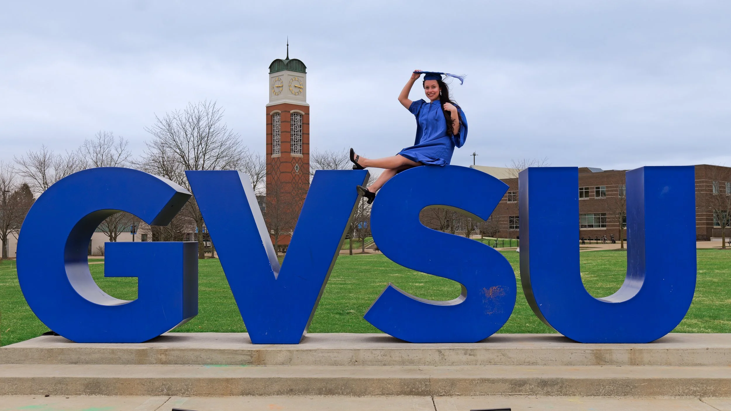 A woman in a blue graduation gown and cap sitting on a large blue 'V' in a park, with a clock tower and leafless trees in the background.