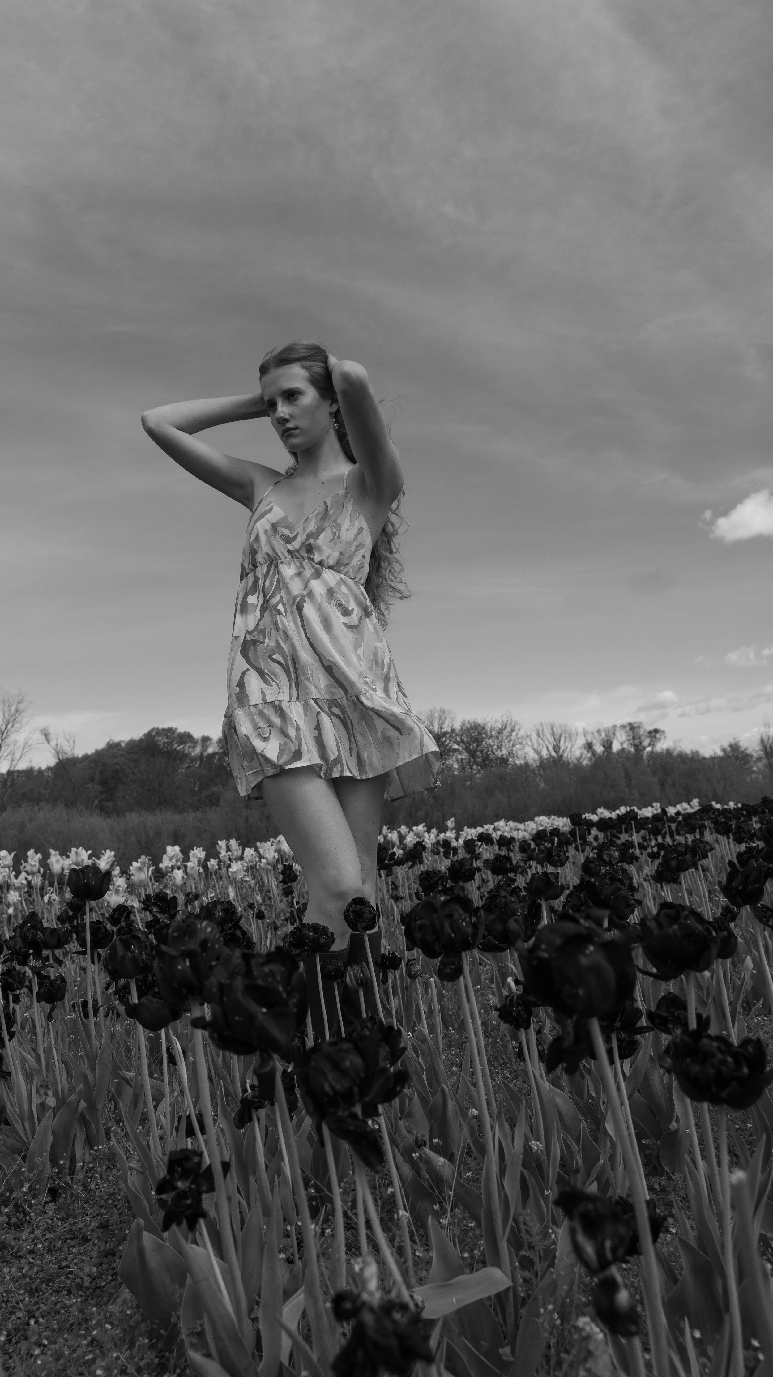 A young woman standing in a field of flowers, holding her hair back with both hands, wearing a patterned dress, under a cloudy sky.