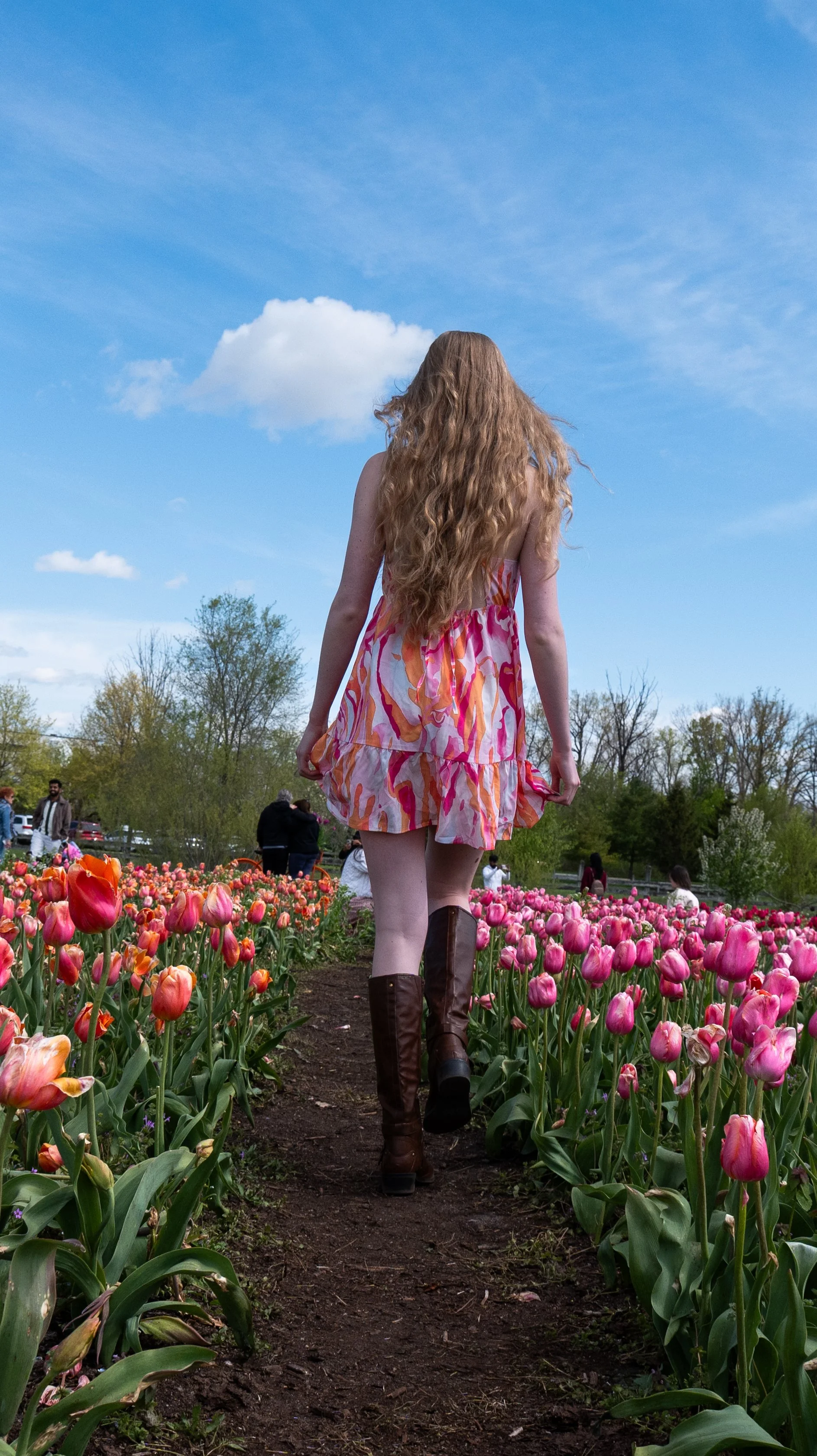 A woman with long red hair wearing a colorful dress and brown boots walking through a tulip garden with other people in the background under a partly cloudy sky.