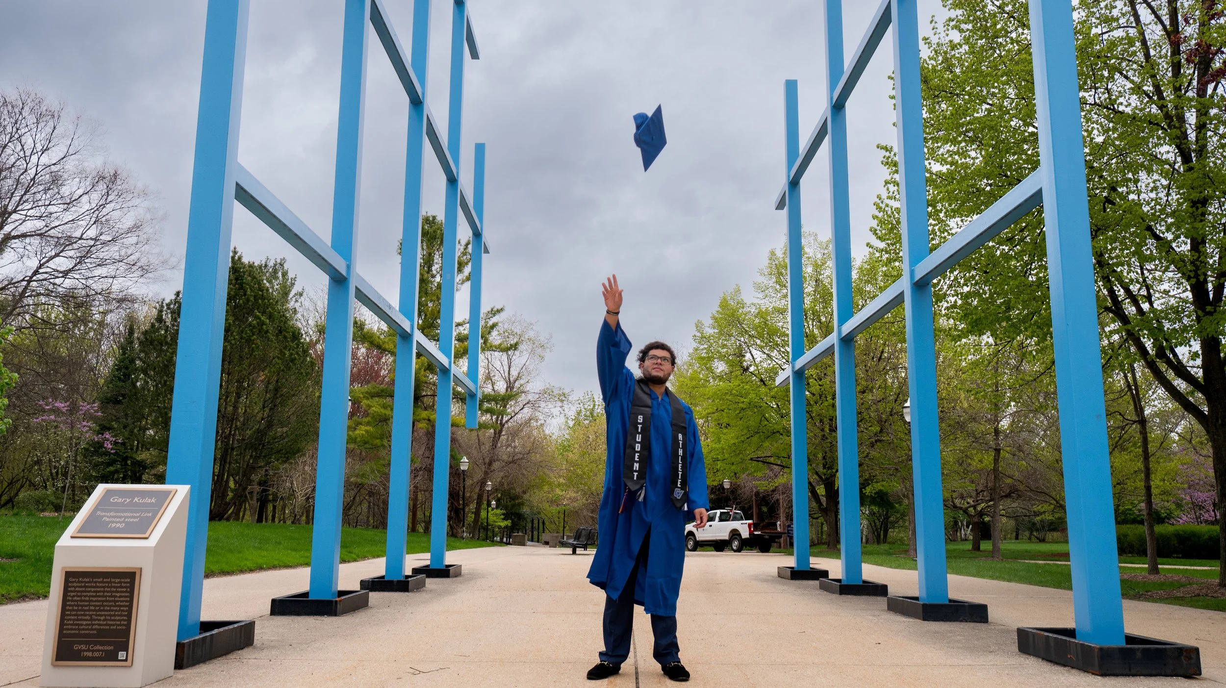 A man in a graduation cap and gown stands under a large blue metal structure, tossing his cap in the air, during a graduation ceremony outdoors.